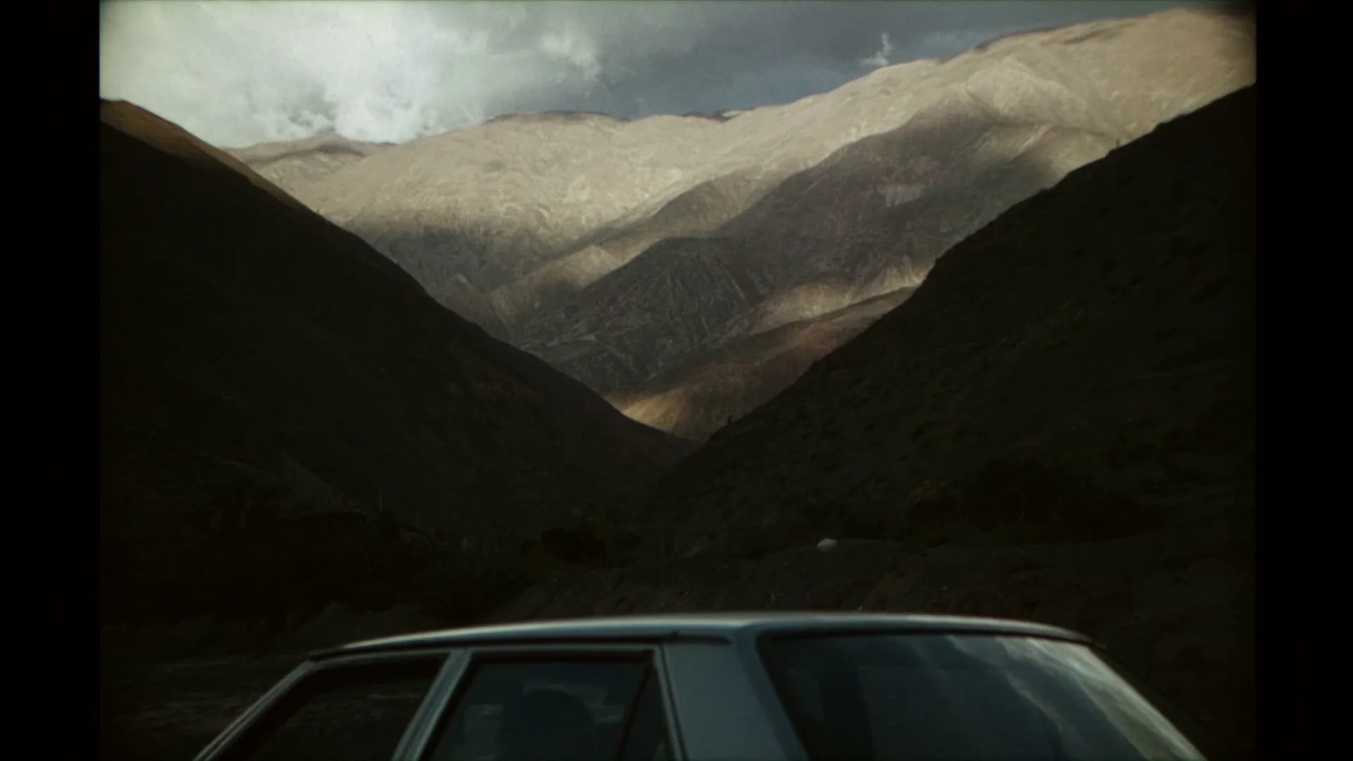 A car is parked in a valley, with towering mountains on either side, some illuminated by sunlight under a cloudy sky.