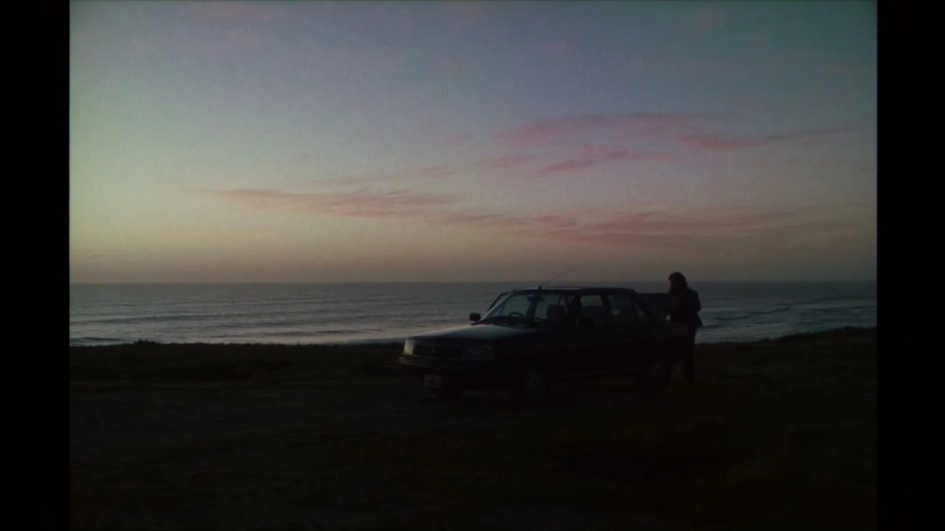 A dark car is parked on a cliff overlooking the ocean at dusk, with a person standing next to the car.