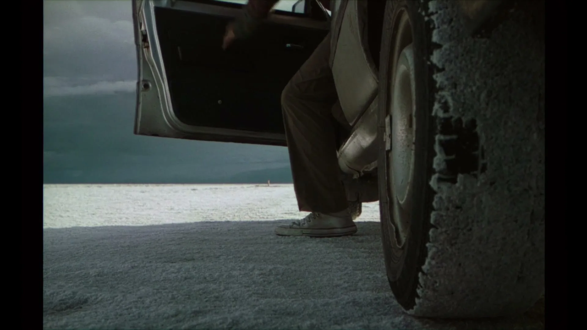 A person in brown pants and white sneakers steps out of a silver car onto a vast, flat, white salt plain under a cloudy sky.