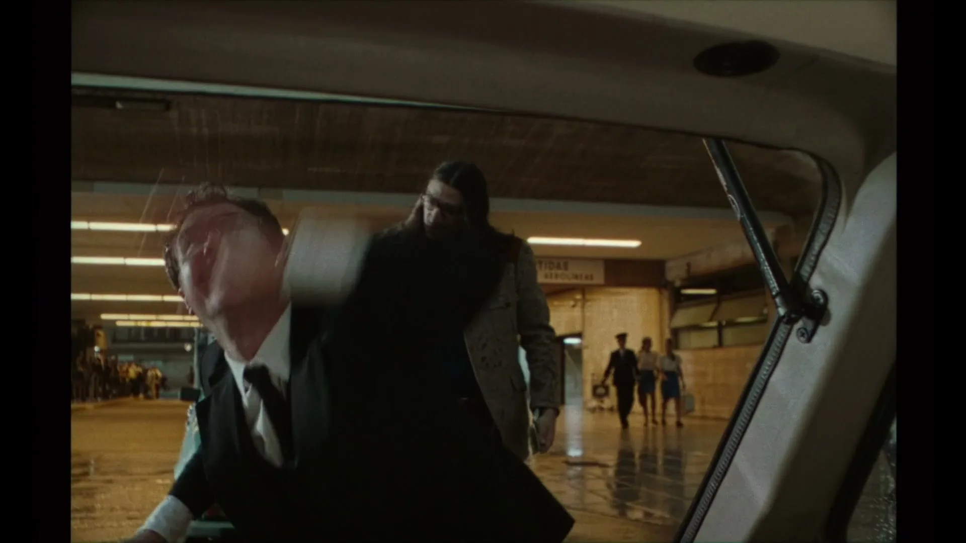 A man in a suit gets into a car, looking back at a long-haired man in sunglasses as rain splashes on the car window in an underground station.