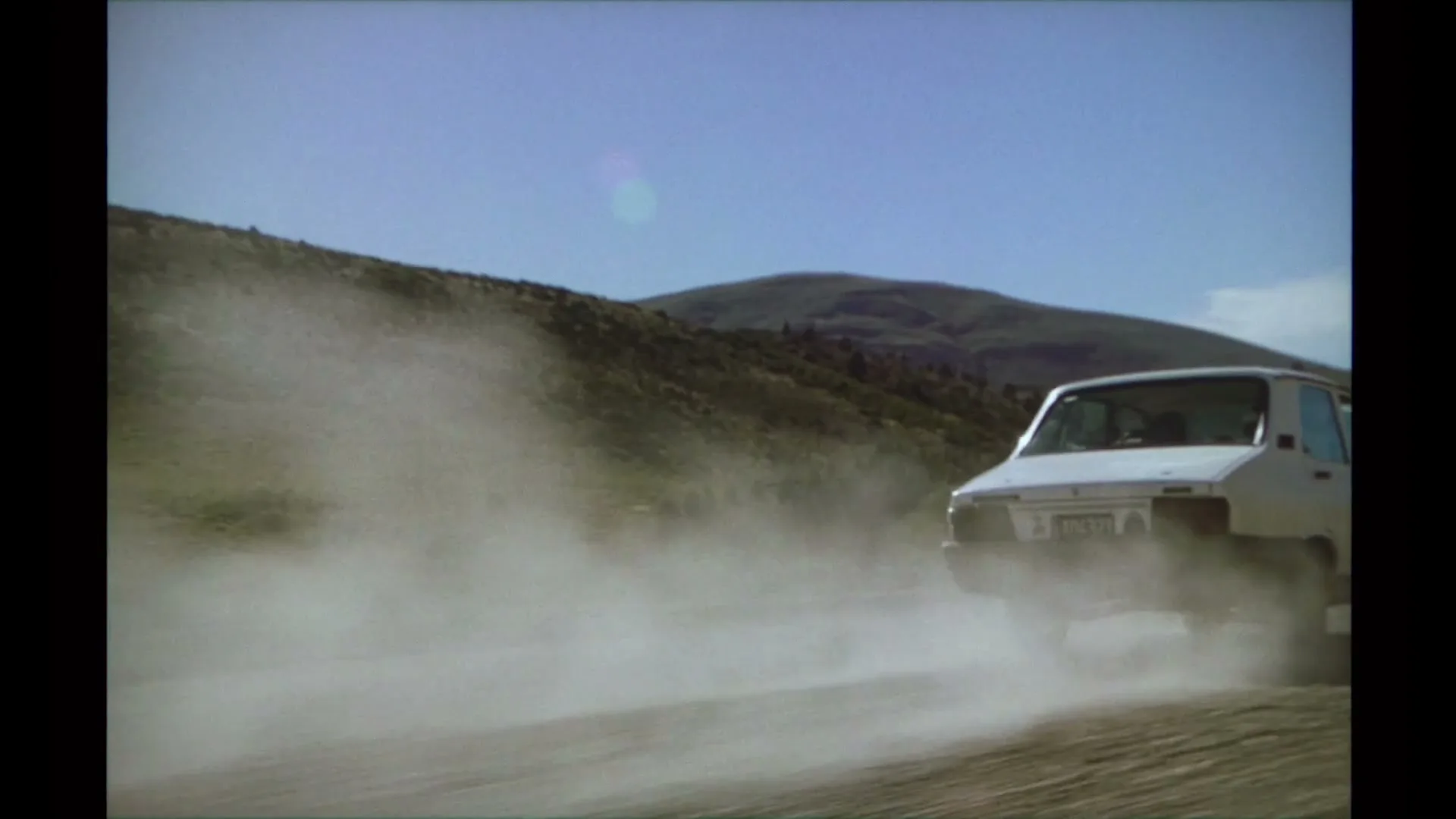 A white car drives away from the camera on a dirt road, kicking up a large cloud of dust in a mountainous, arid landscape.