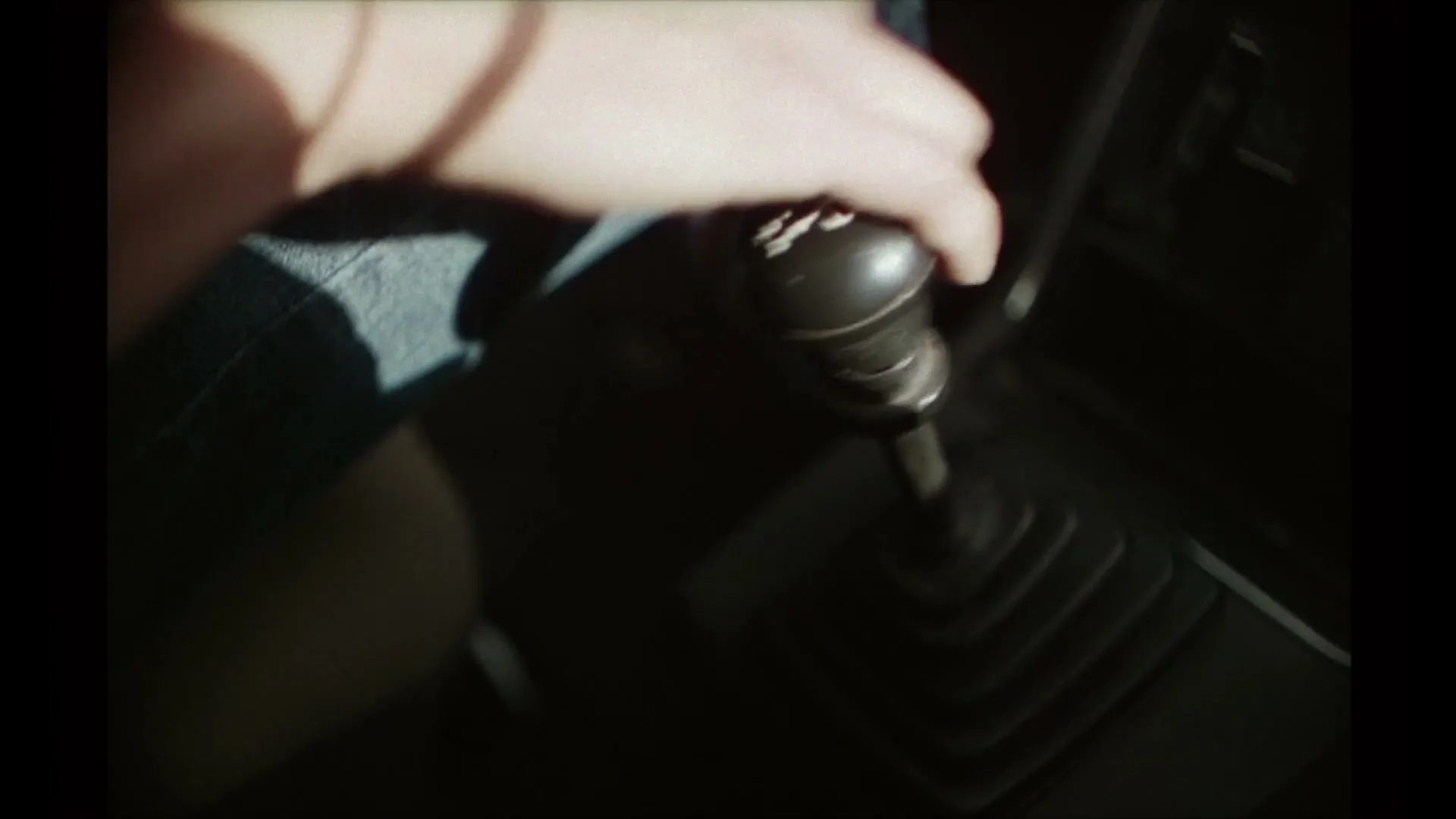 A person's hand, wearing a blue denim shirt, shifts gears in a car, with sunlight casting shadows inside the vehicle.
