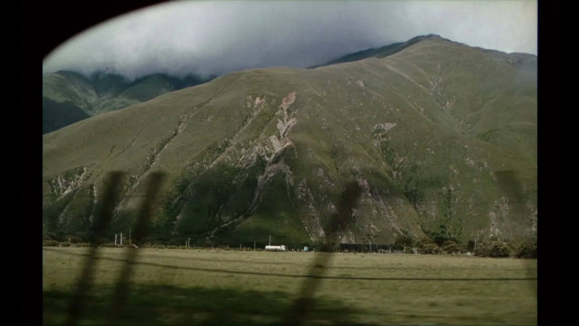 A wide shot from inside a moving vehicle shows a vast green mountain range under a cloudy sky, with a field in the foreground.
