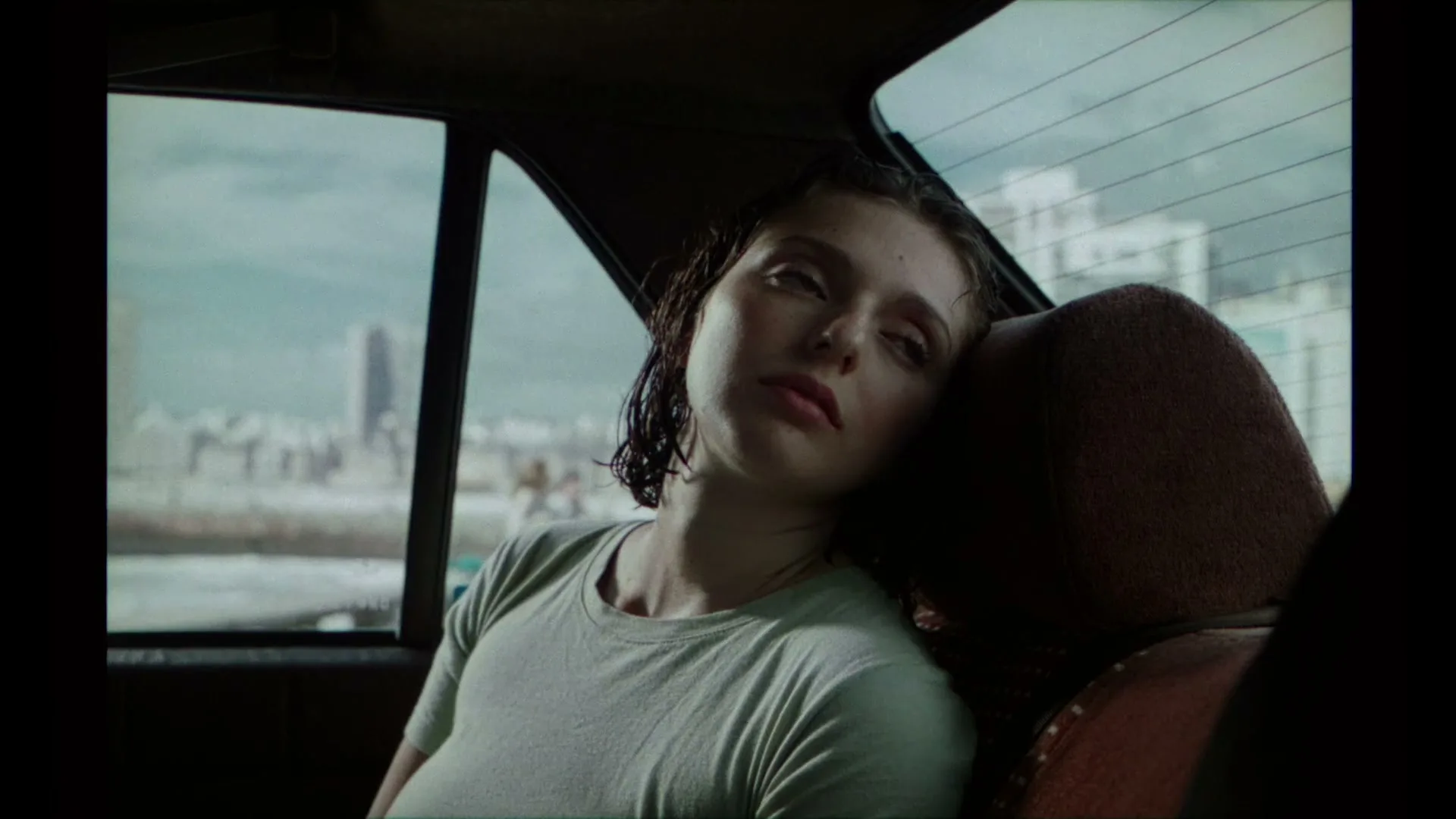 A young woman with wet hair, wearing a light green t-shirt, sleeps in the back seat of a car, with a blurry cityscape visible outside the window.