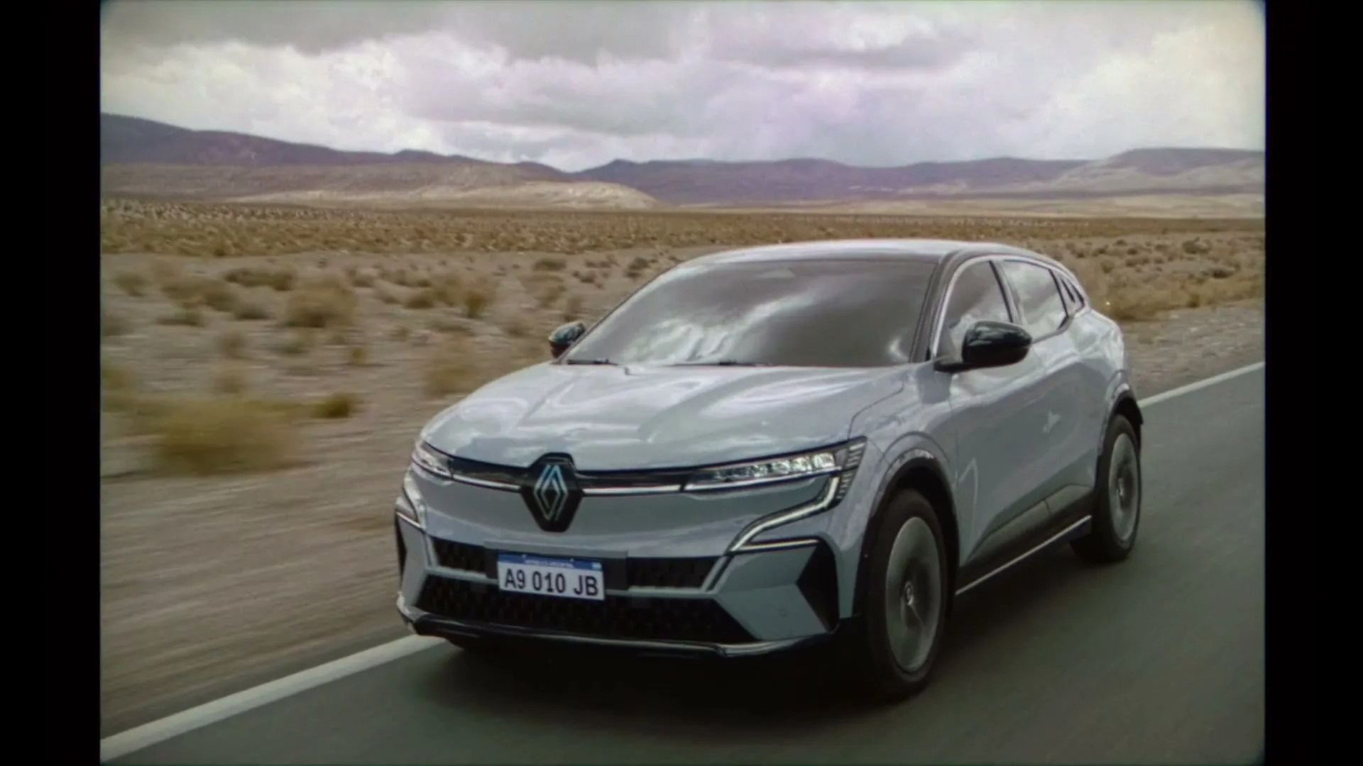 A light grey Renault car drives on a desert road under a cloudy sky, with mountains in the background.