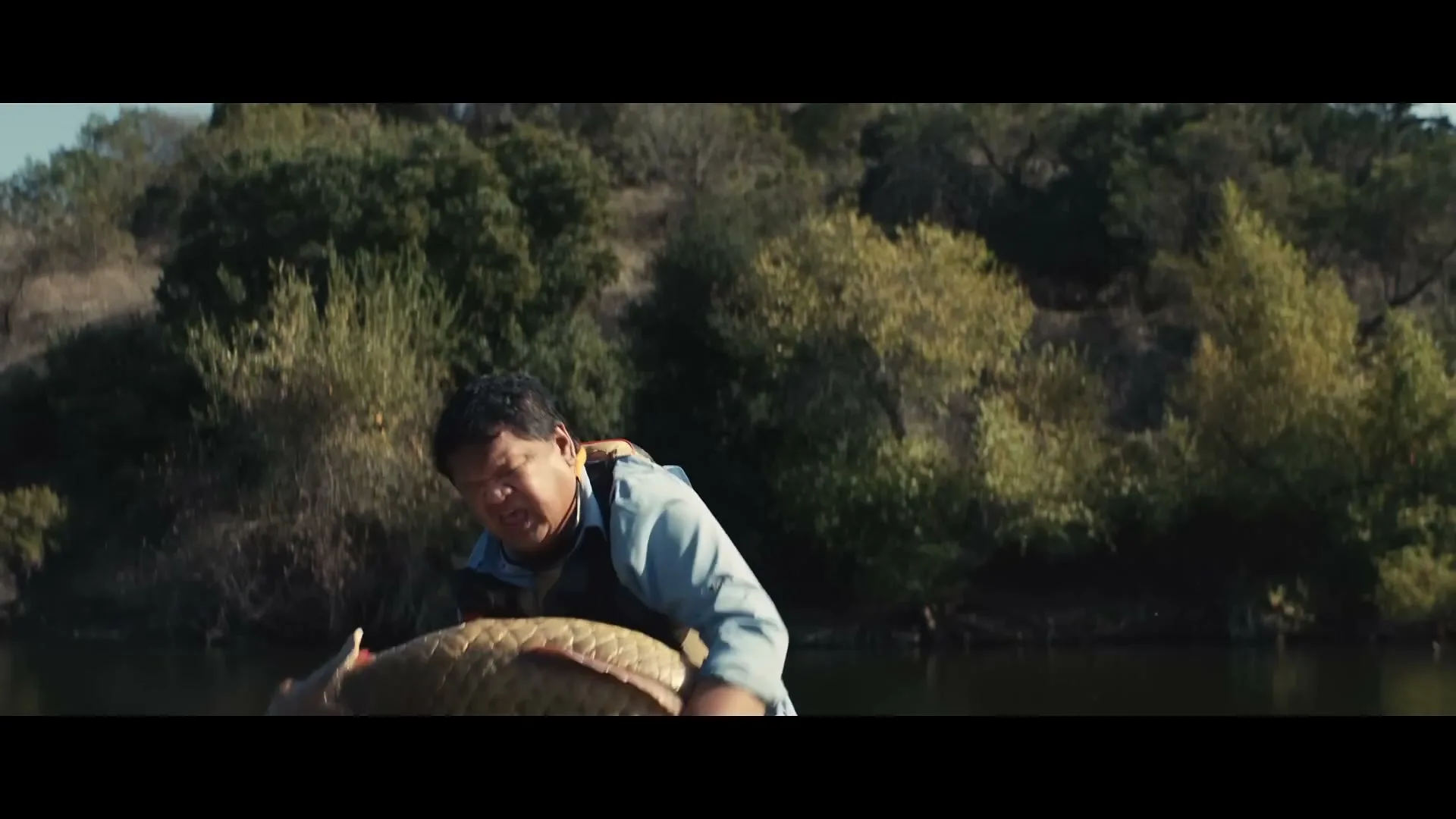 A man in a boat struggles to hold a giant fish, while a smartphone on the boat records him, with trees in the background.