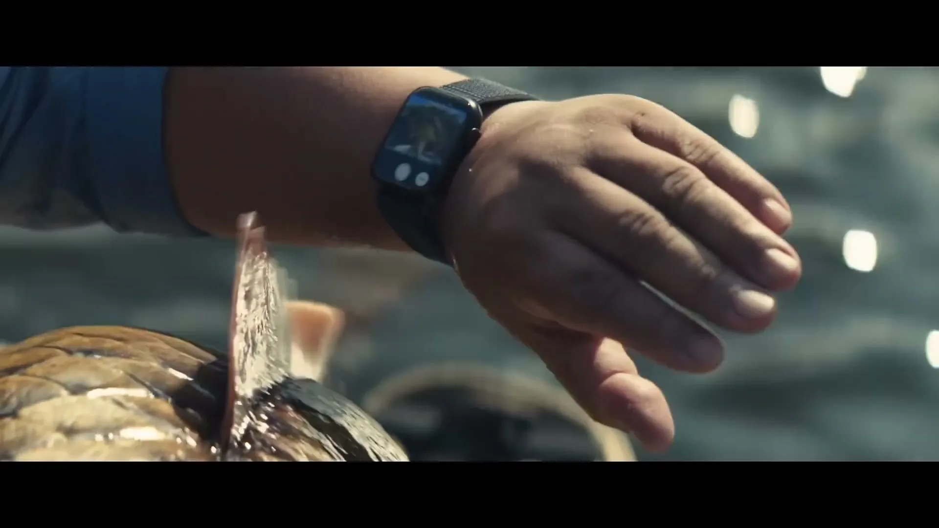 A man's arm and hand are shown wearing an Apple Watch, holding a fish with a hook in its mouth, with water in the background.
