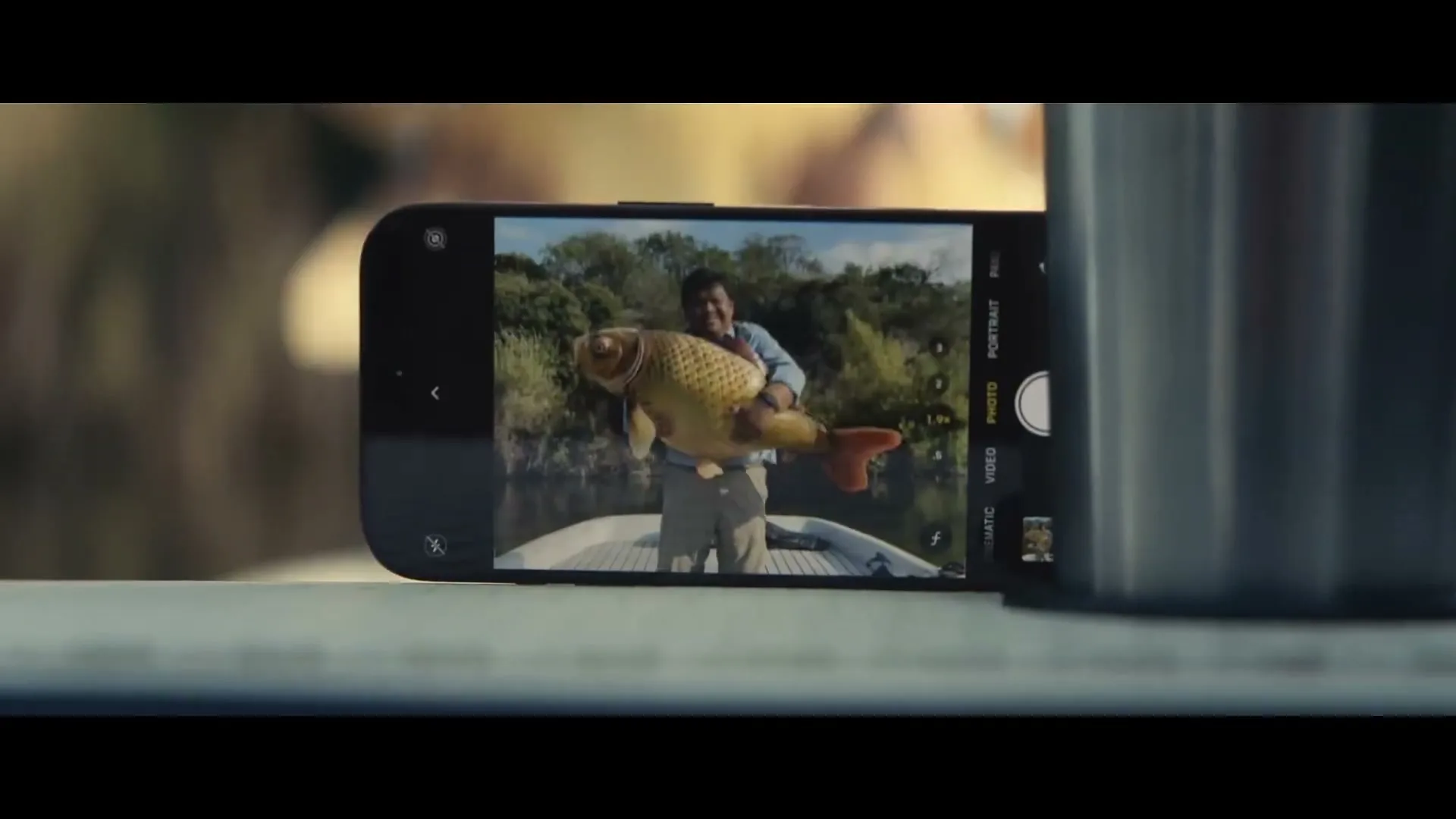 A close-up shot of an iPhone screen shows a man holding a large fish on a boat, with a blurred background of trees and water.