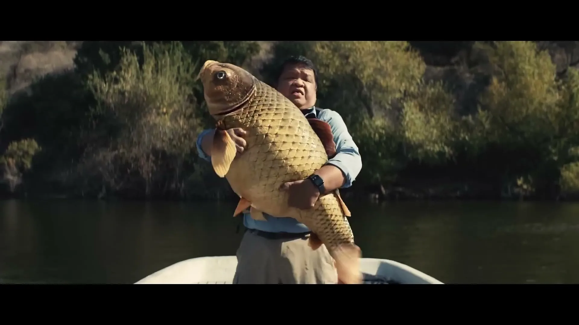 A man in a fishing boat struggles to hold a very large fish, with a tree-lined shore in the background.