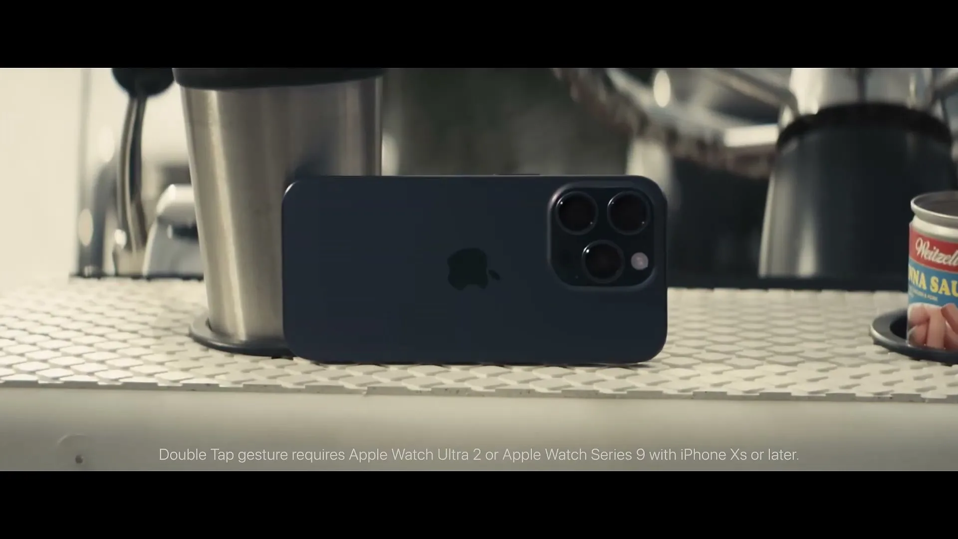 A dark blue iPhone 15 Pro Max lies on a textured surface next to a metal cup and a can of Vienna sausages.