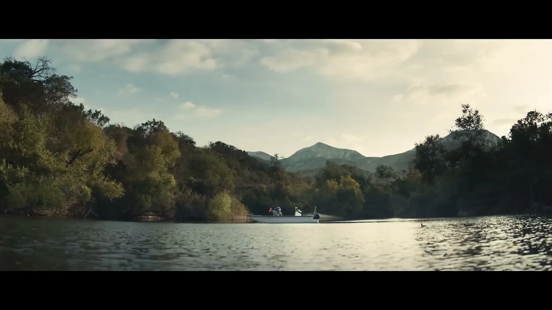 A wide shot of a small boat with two people on a lake, surrounded by green trees and mountains under a cloudy sky.