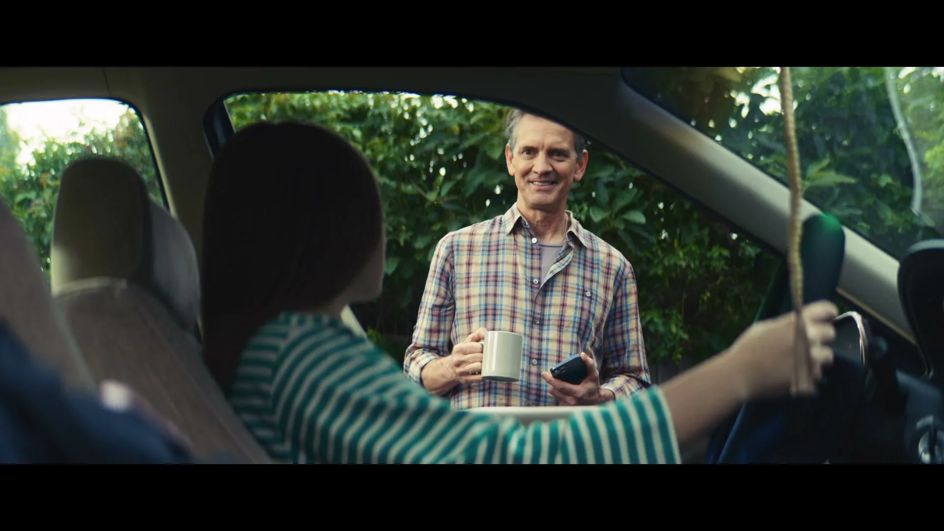 A teenage girl sits in the driver's seat of a car, looking at an older man holding a mug and phone outside the car window.