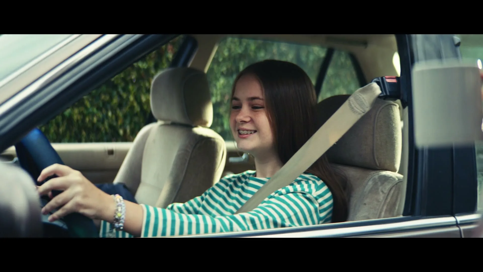 A young female driver with long brown hair and a striped shirt smiles while sitting in a car, looking up and to the right as the car begins to move.