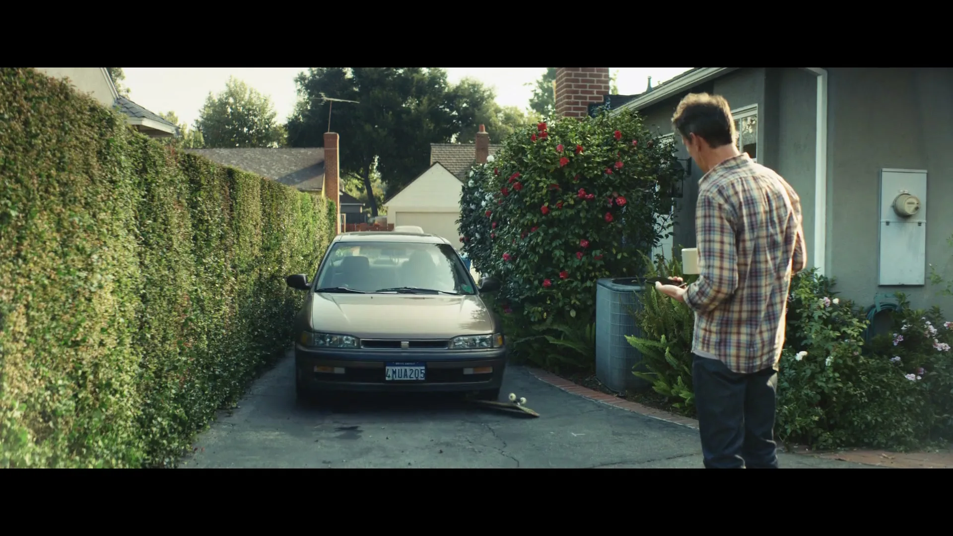 A father stands in a driveway, holding a phone and looking at his daughter in a car, as a skateboard rolls away.