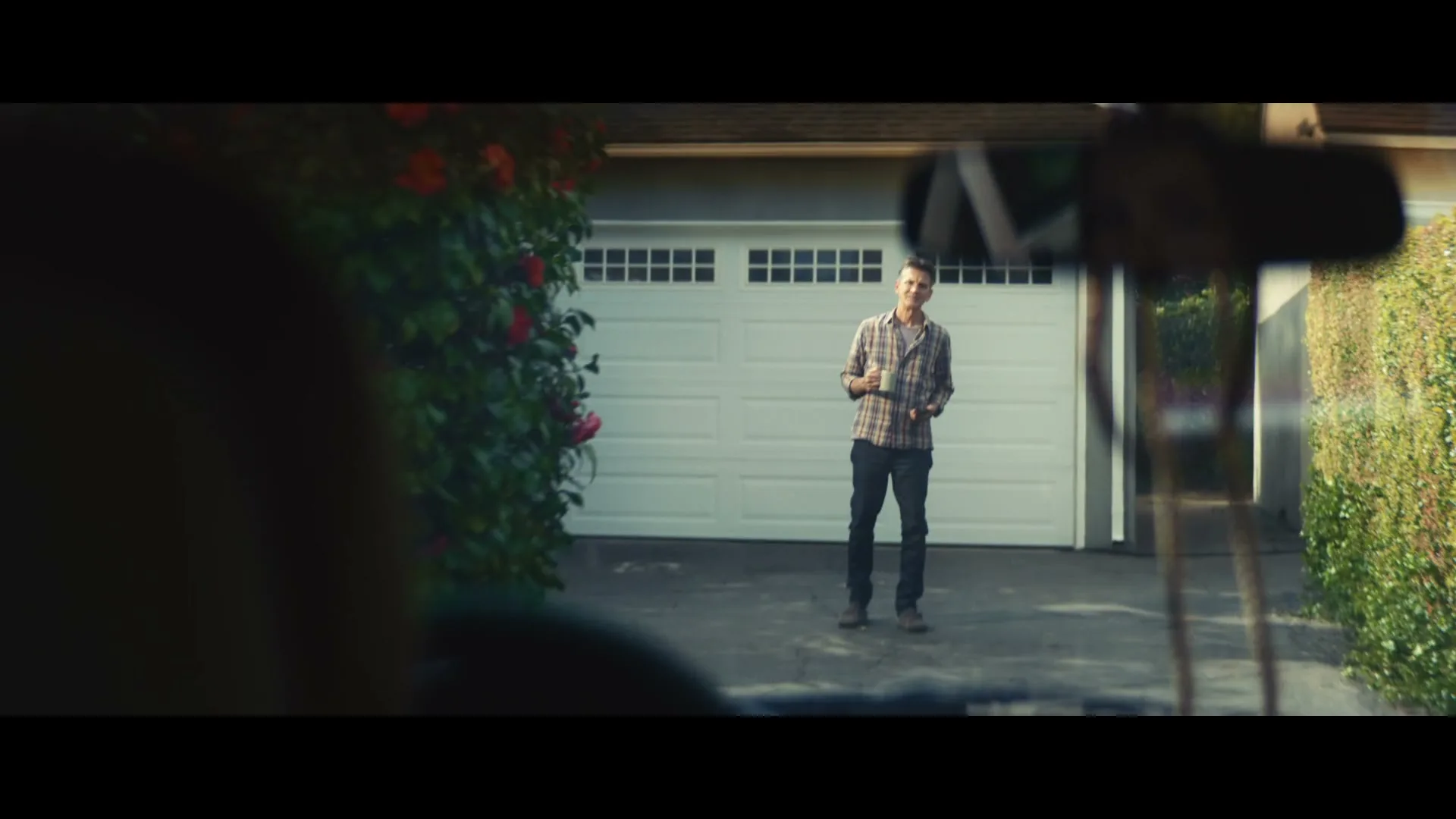 A father, wearing a plaid shirt, stands in a driveway holding a mug and waving goodbye to someone in a car.
