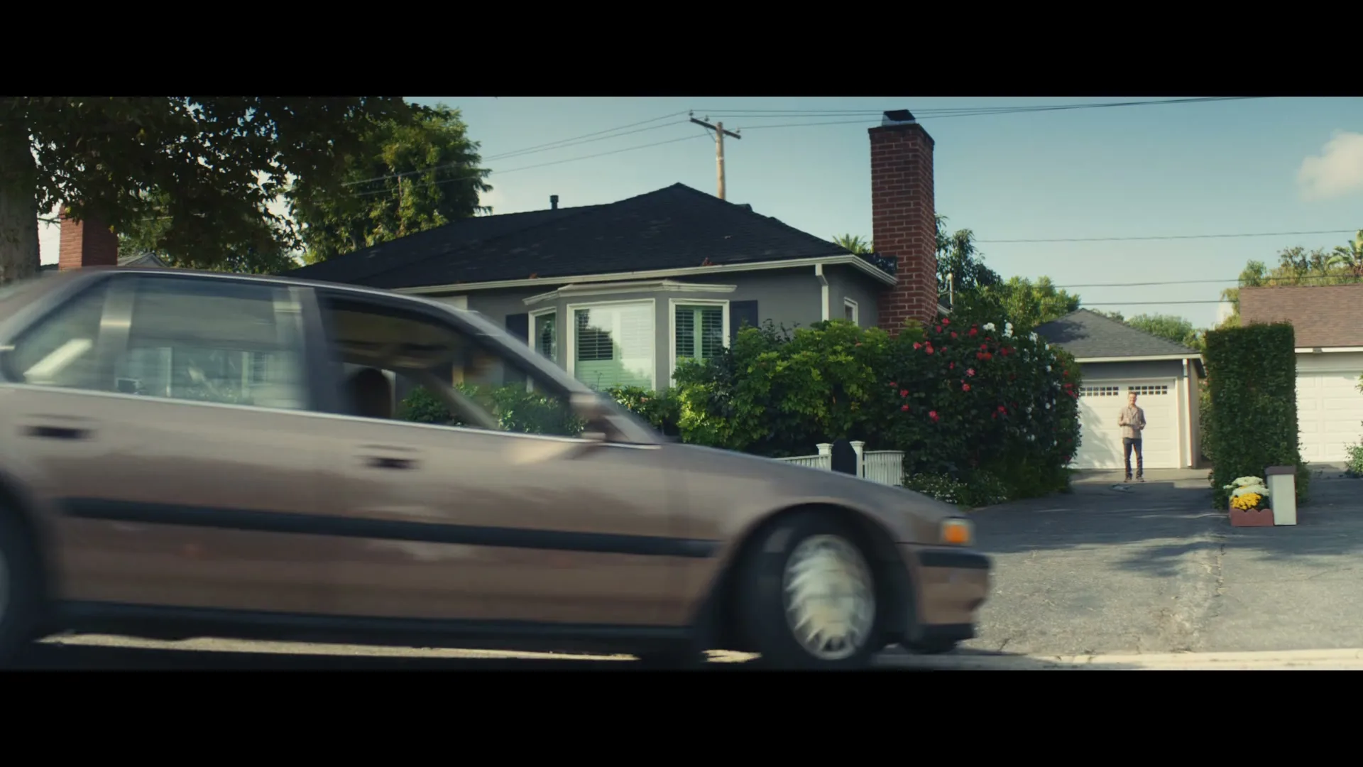 A brown car backs out of a driveway, hitting a blue recycling bin, as a man watches from the garage.