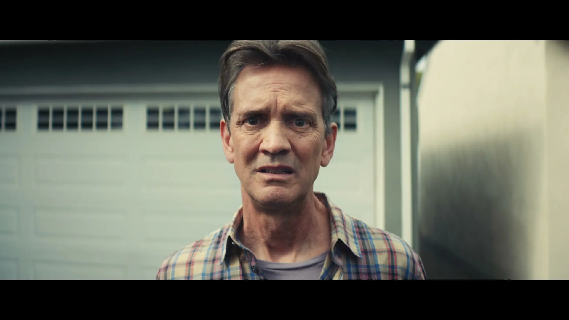 A middle-aged man with a concerned expression stands in front of a garage door, looking directly at the camera.