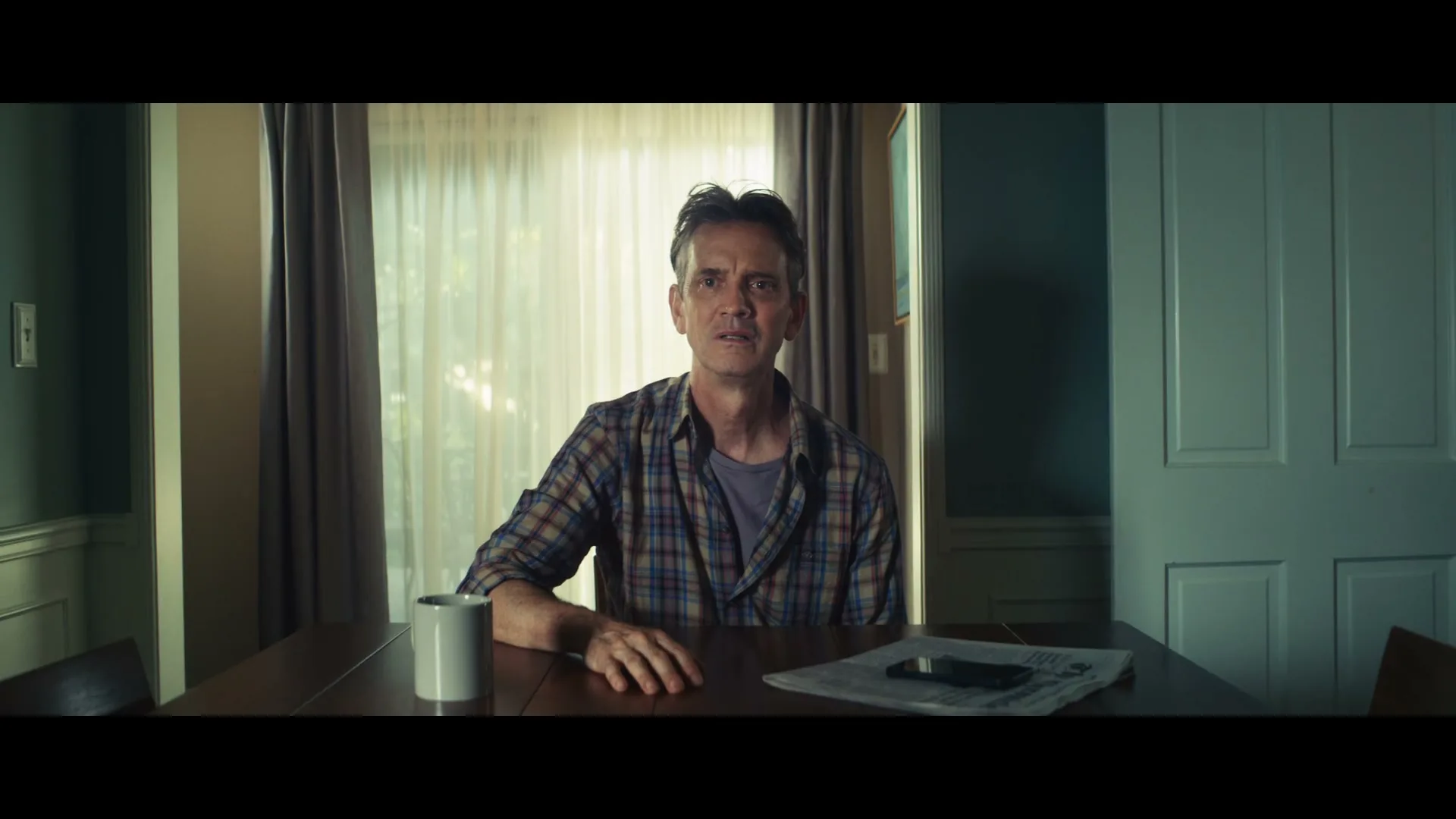A middle-aged man with a plaid shirt sits at a dining table, looking concerned, in a suburban home during the day.