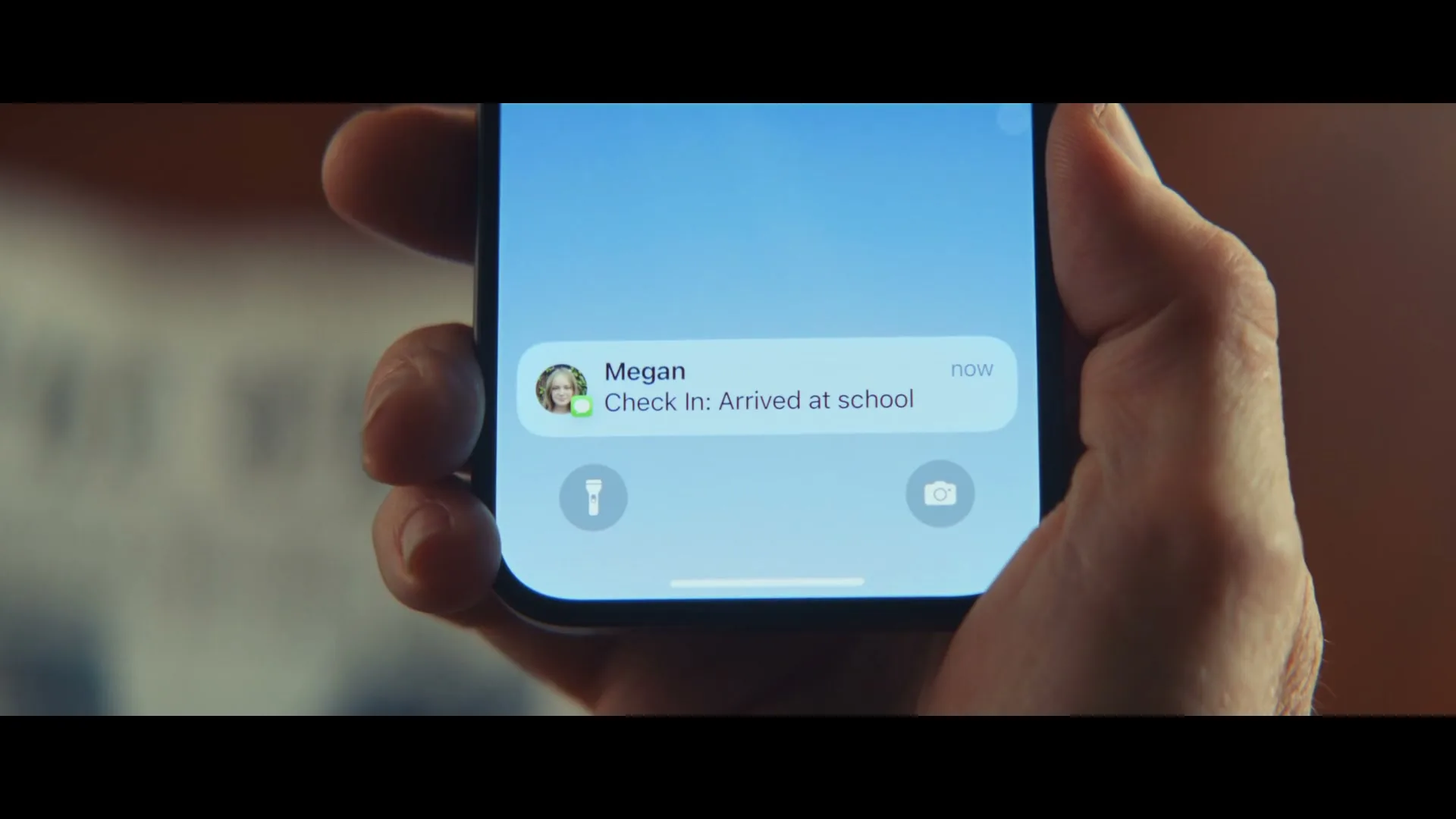 A person's hand holds an iPhone, displaying a "Check In: Arrived at school" notification from "Megan" on a light blue screen.