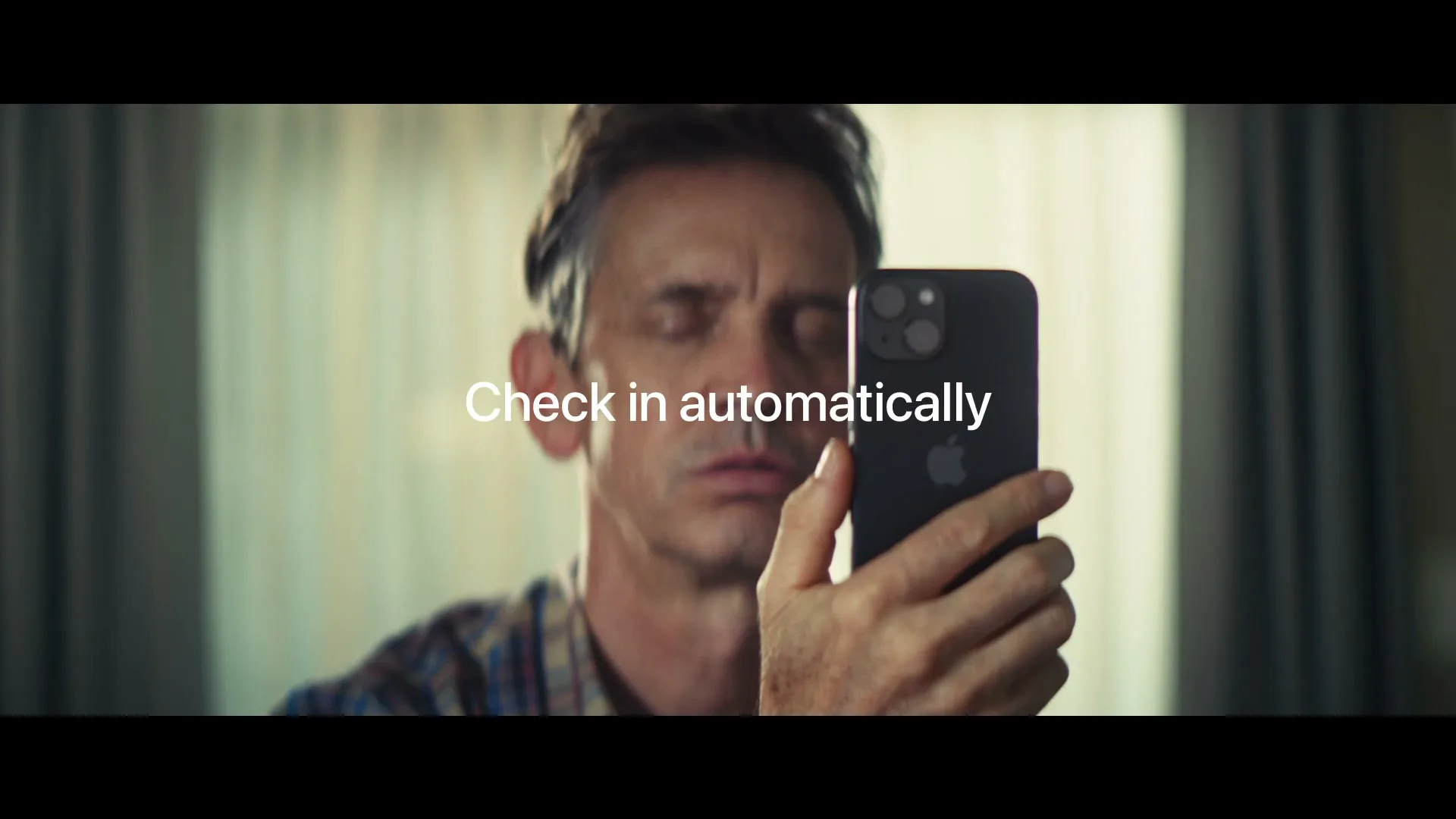 A middle-aged man with a worried expression holds up a black iPhone, looking at it intently, in a softly lit interior.