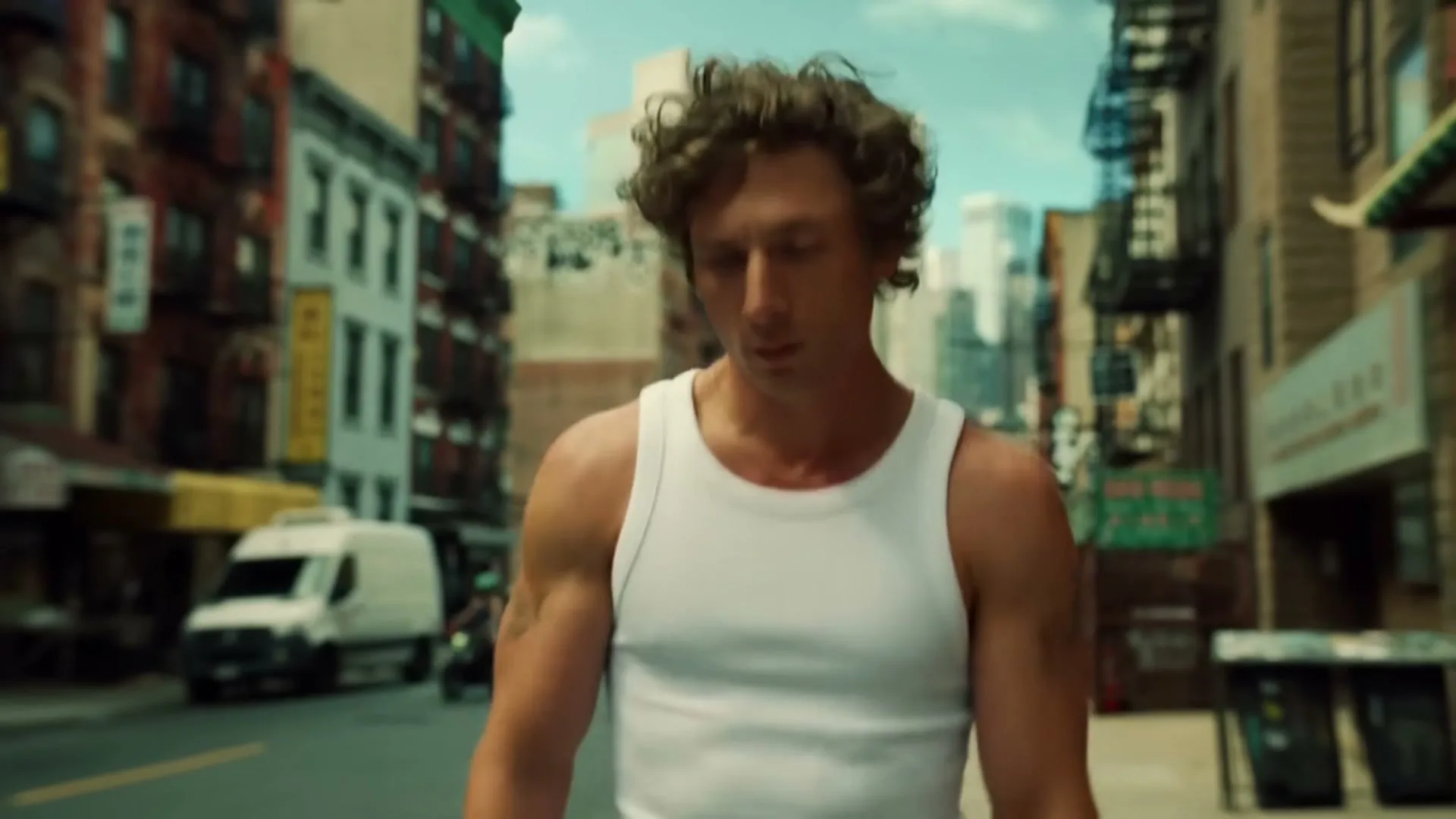A man with curly hair in a white tank top walks down a city street, looking around.