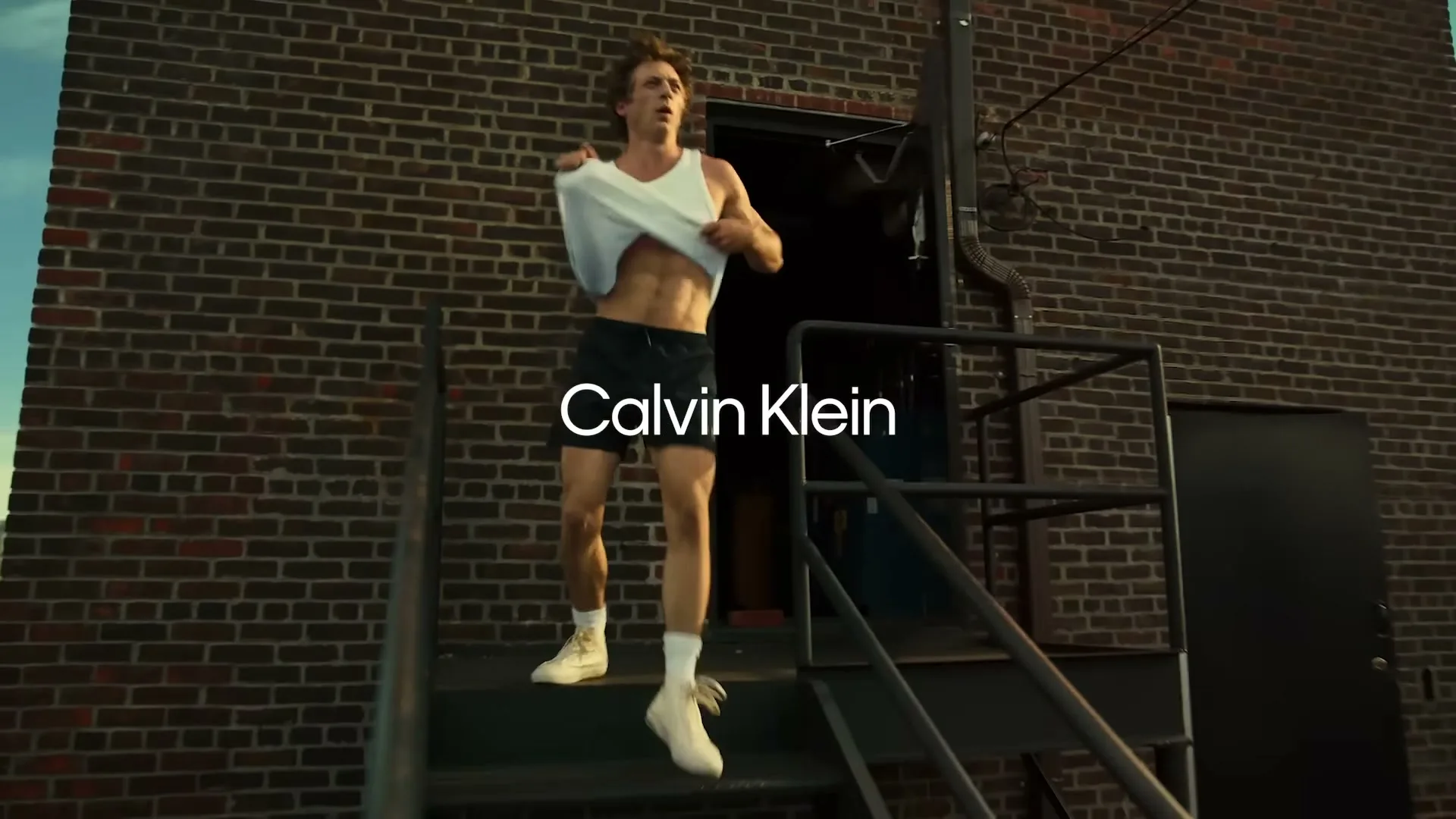 Jeremy Allen White, a man in his 30s, runs up a metal staircase on a brick building, taking off his white tank top.