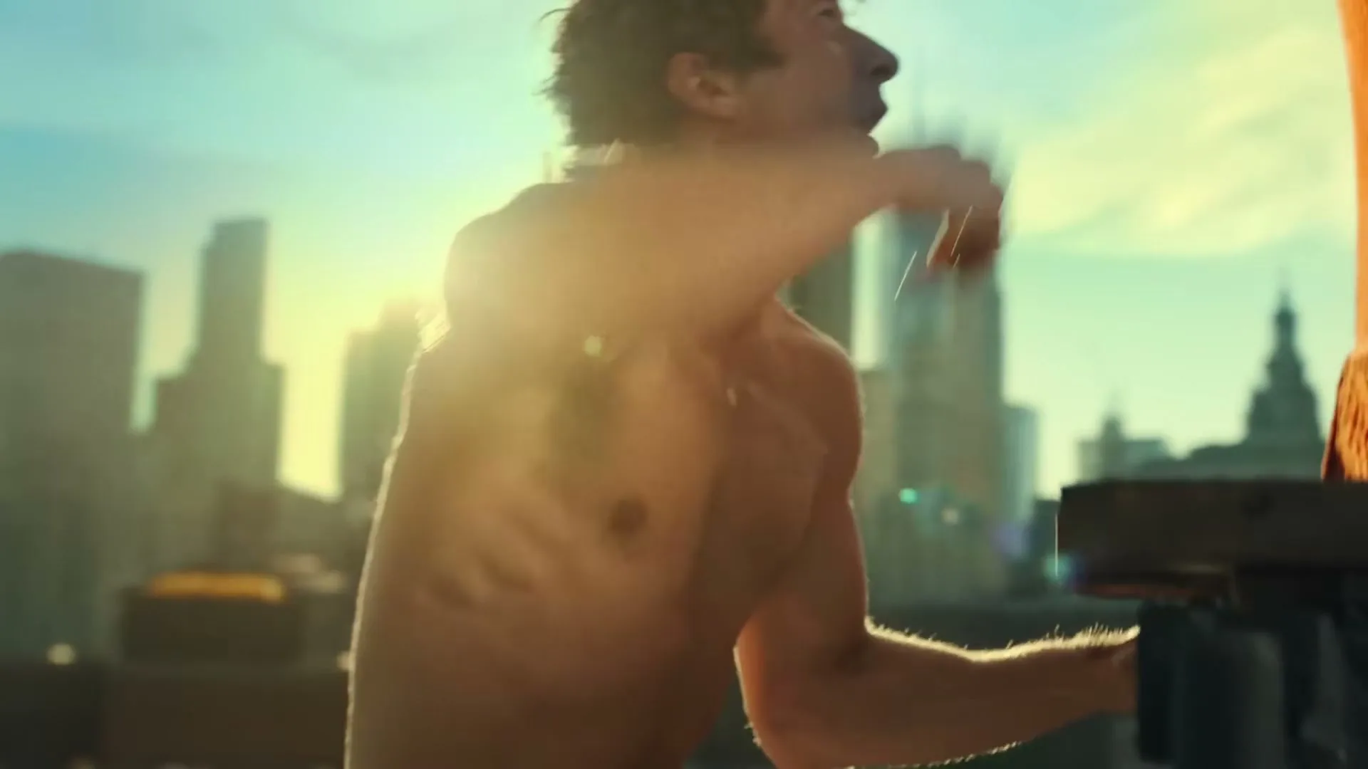 A shirtless man with curly hair stretches his arm up against an orange cushion on a rooftop with a city skyline in the background.