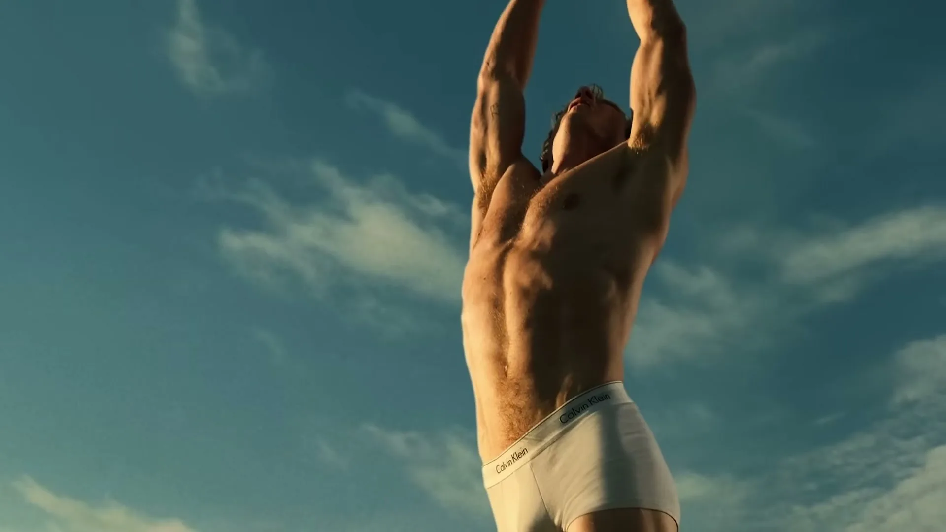 A muscular man in white Calvin Klein underwear stretches his arms above his head against a blue sky with clouds.