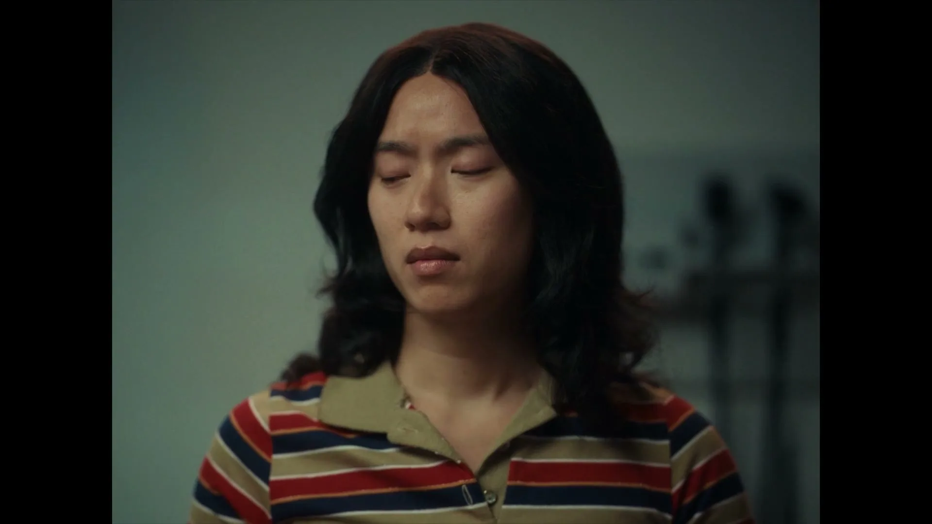 A young person with dark hair and a striped shirt looks down with a pensive expression in an interior setting.