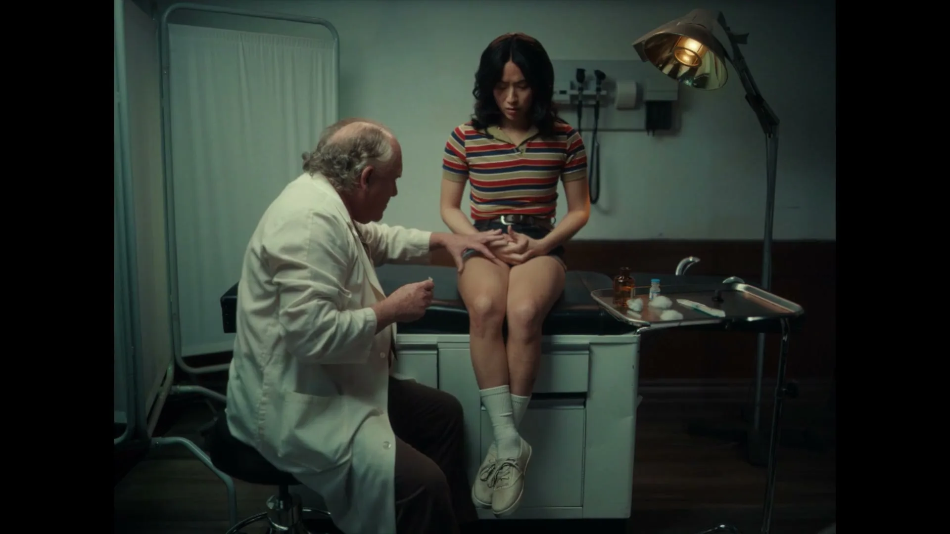 A male doctor in a white lab coat prepares to give an injection to a young female patient sitting on an examination table in a dimly lit doctor's office.
