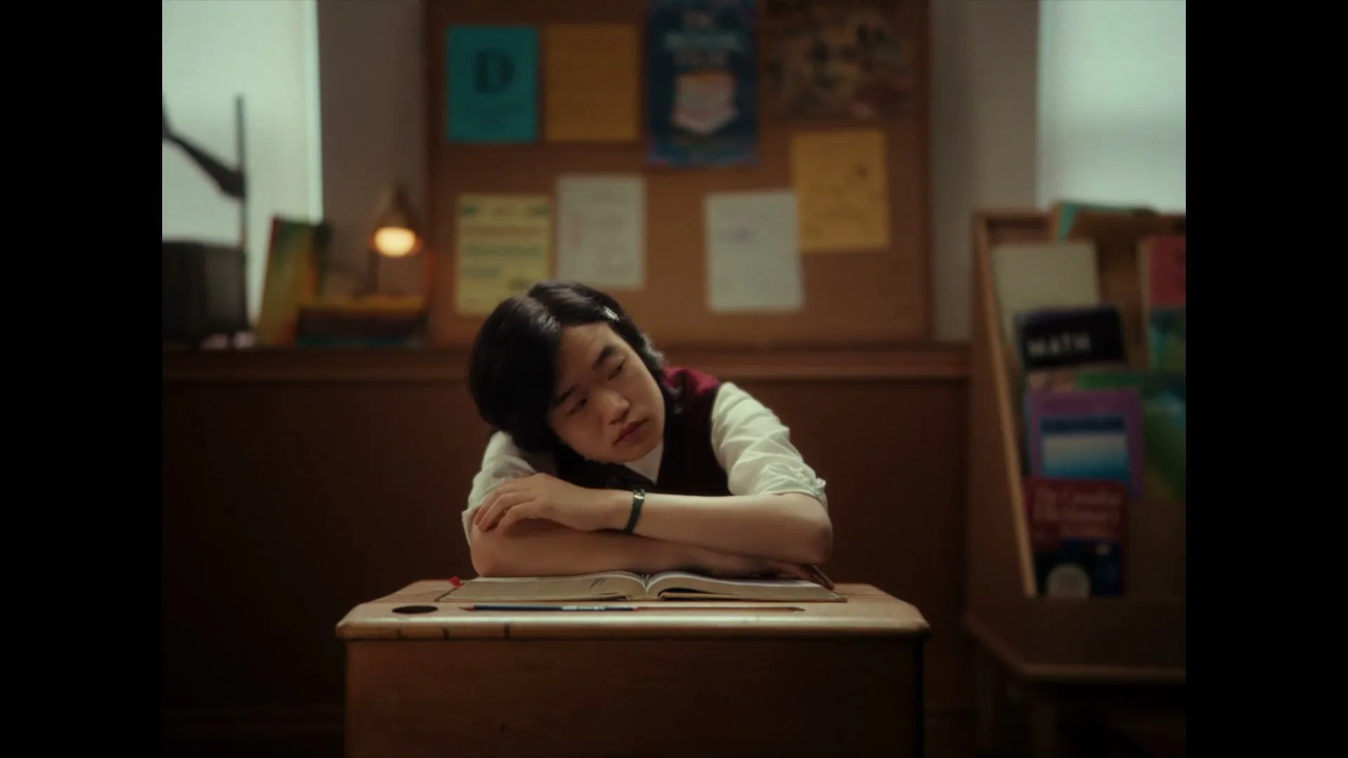 A young person with dark hair and a white shirt with a maroon vest sits at a wooden desk, looking bored in a classroom.
