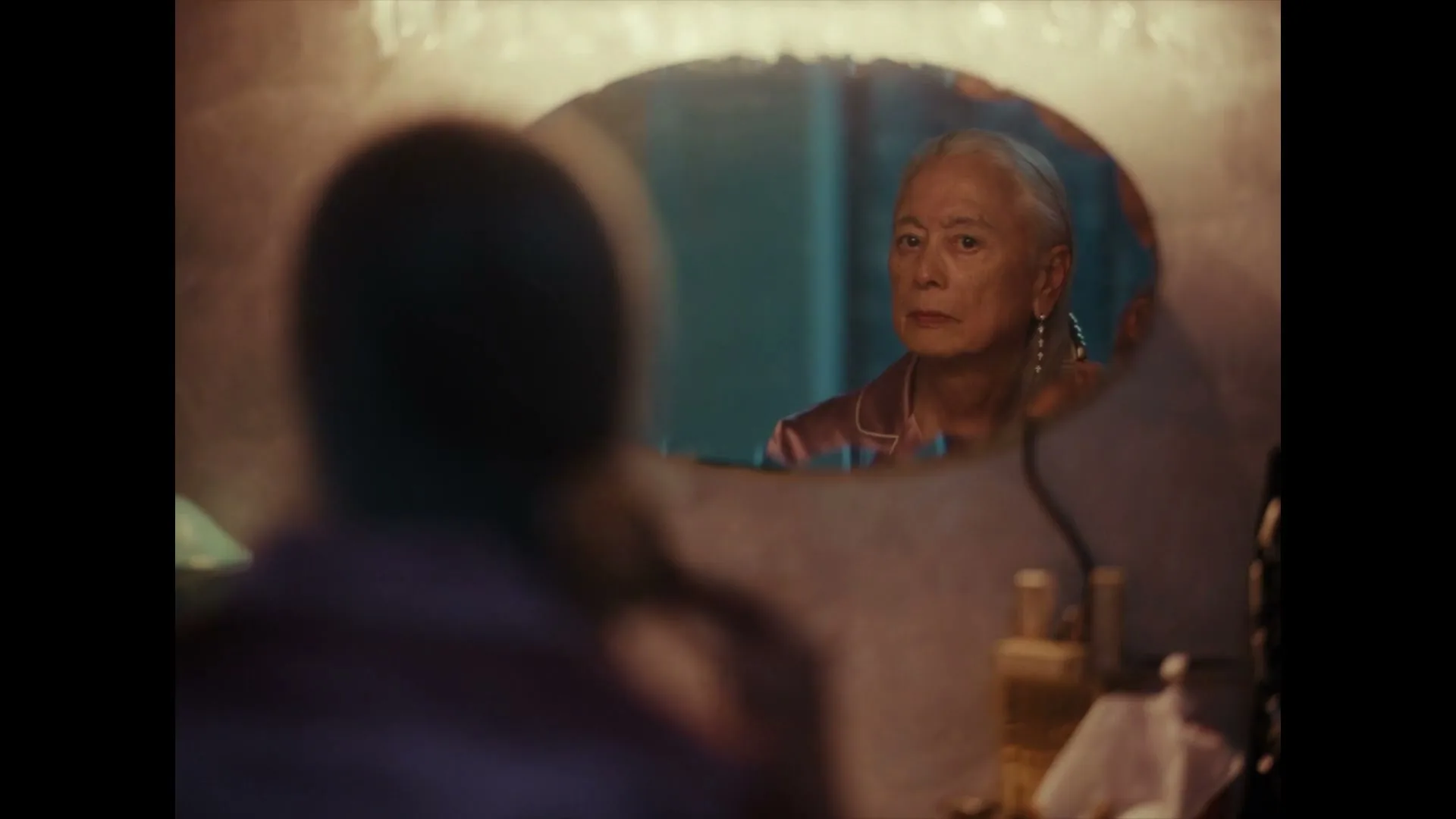 An older person with gray hair looks at their reflection in a mirror, brushing their hair, in a dimly lit room.