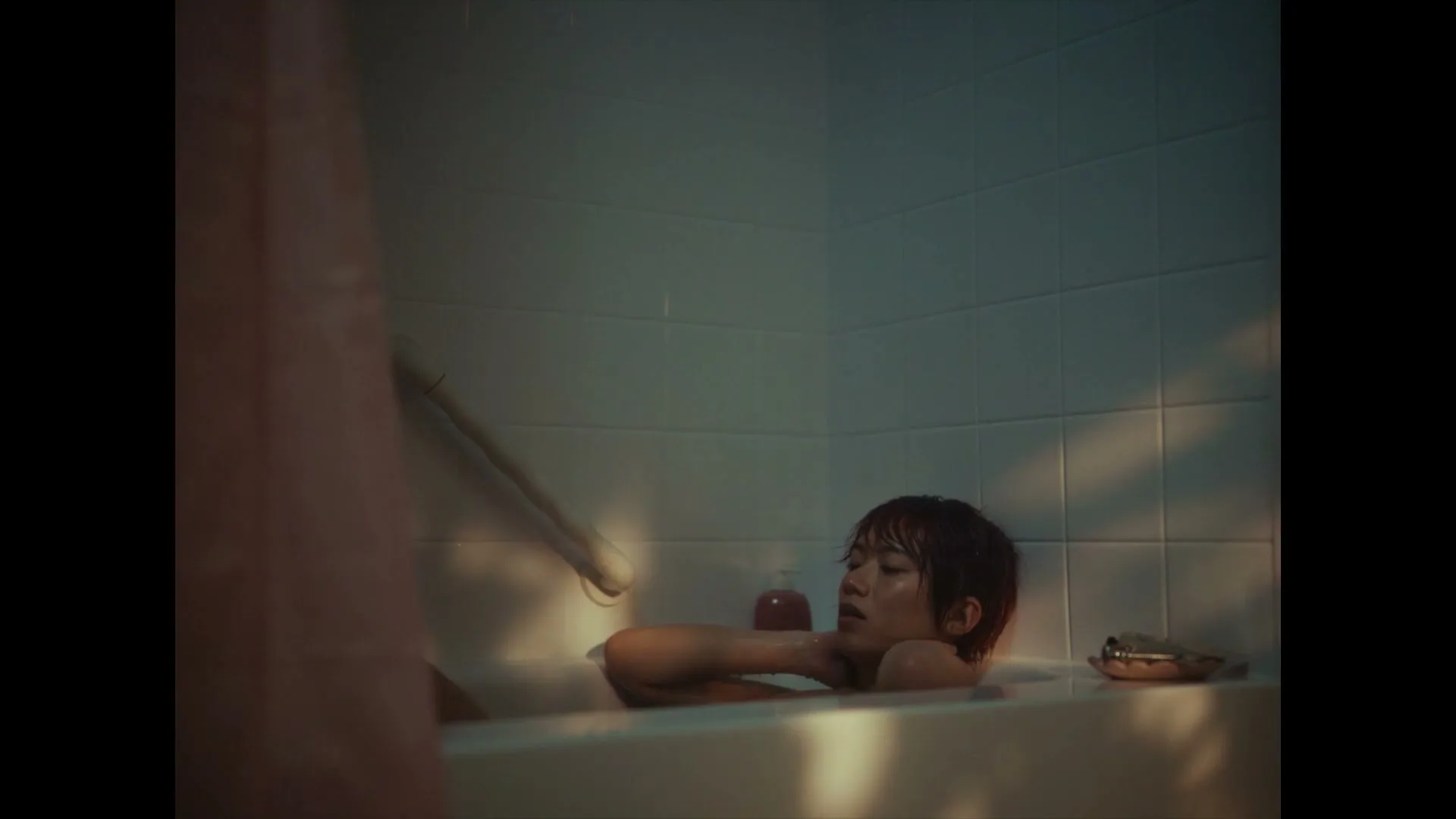 A young person with short hair is seen from the chest up, lying in a bathtub, looking up with a worried expression.