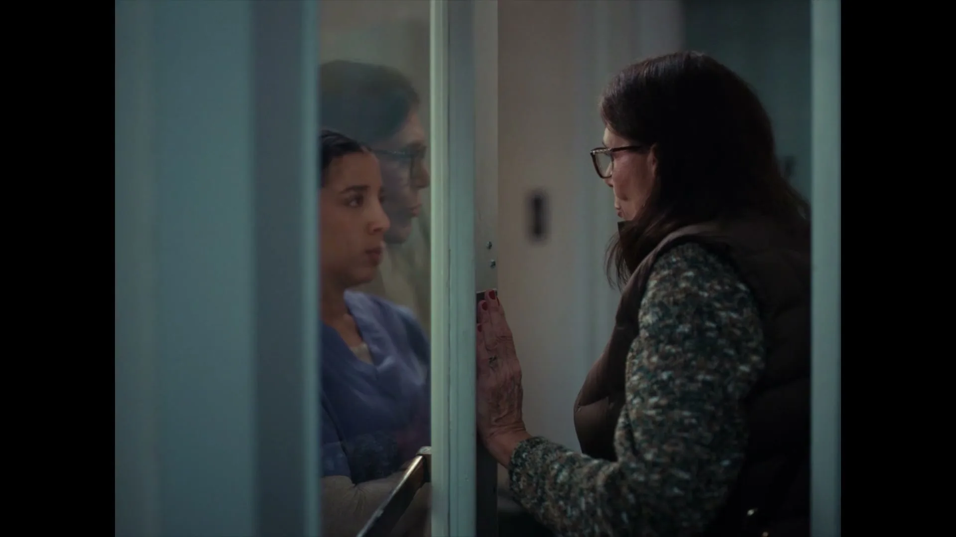 A concerned older woman with dark hair and glasses presses her hand against a glass door, looking at a younger woman in scrubs on the other side, in a hospital hallway.