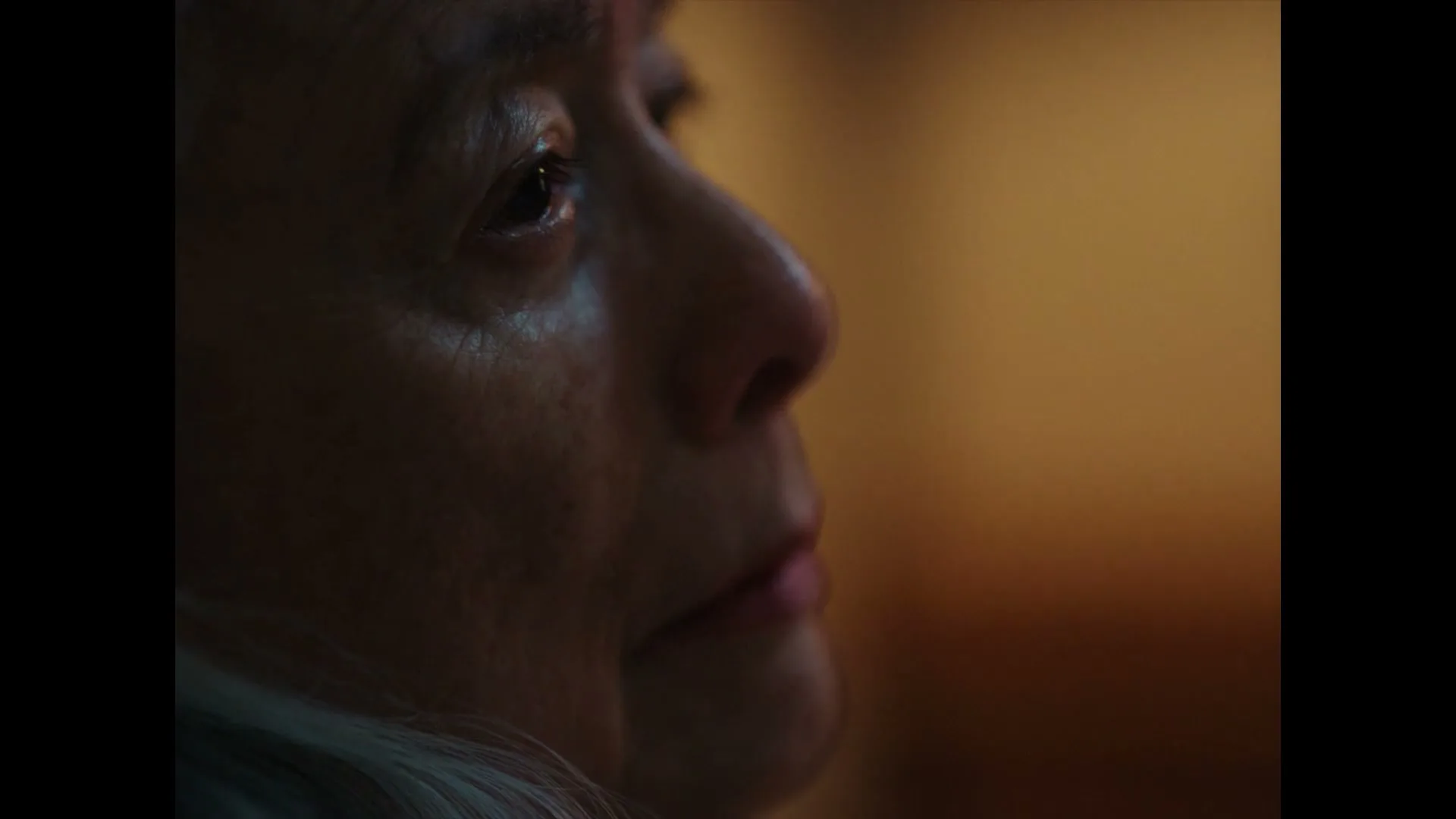 A close-up of an older person's face, looking off-screen with a neutral expression in a dimly lit interior.