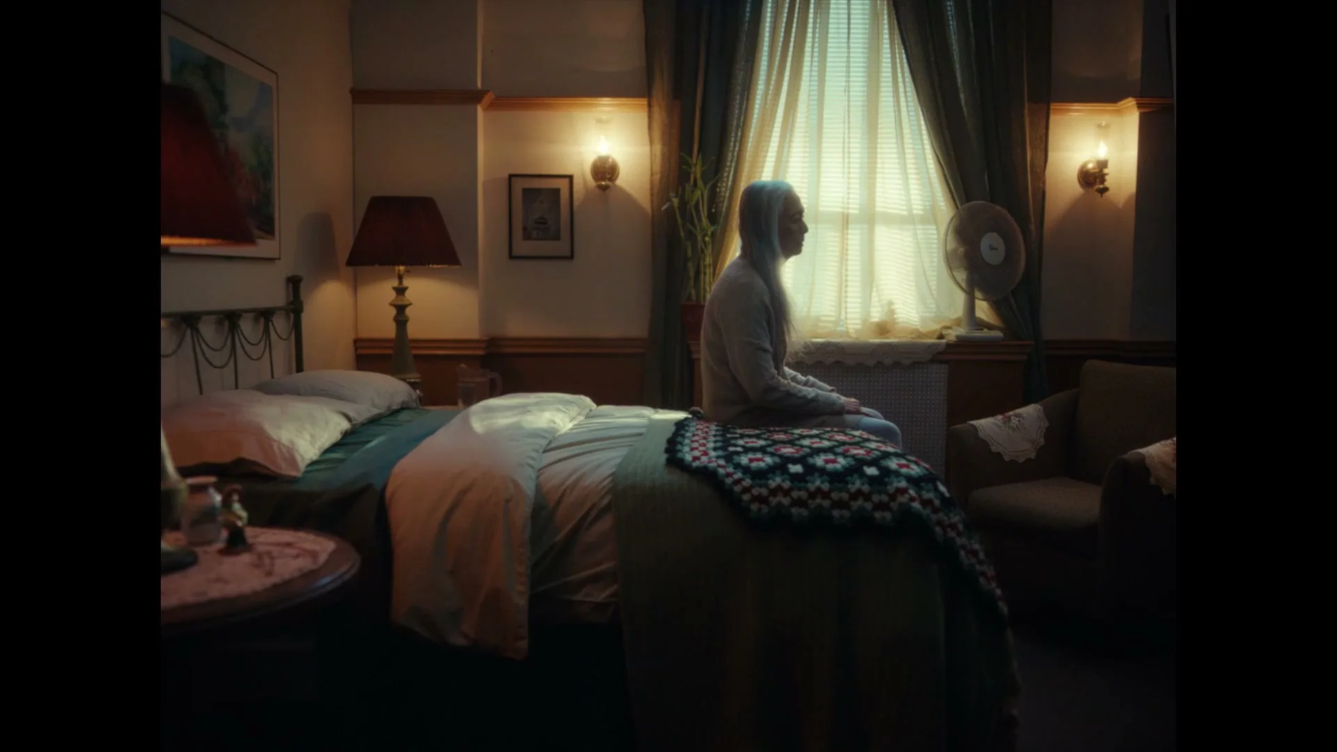 A woman with long white hair sits on a bed, looking out a window in a dimly lit room.