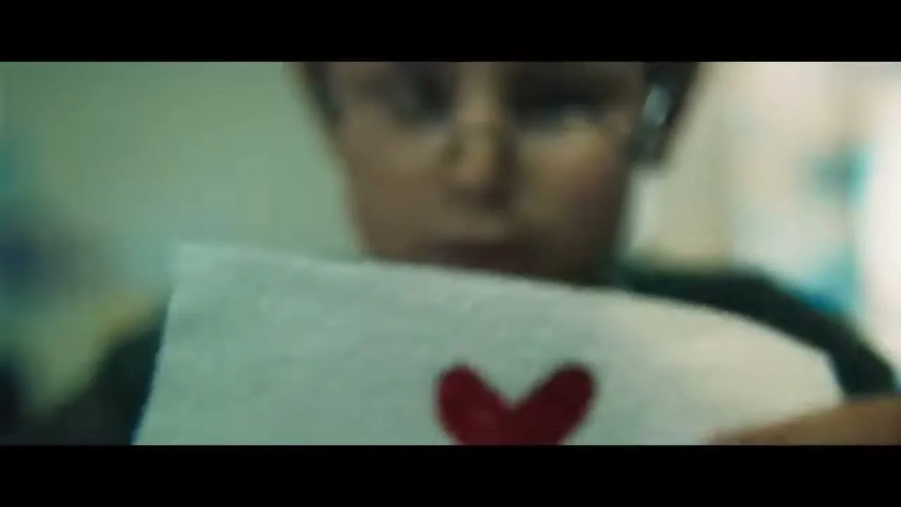 A young boy with glasses holds up a piece of paper with a red heart drawn on it, looking at it intently.