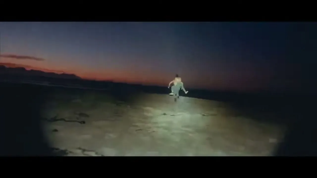 A young woman in a light-colored dress runs away from the camera on a dark beach at dusk, illuminated by a spotlight.