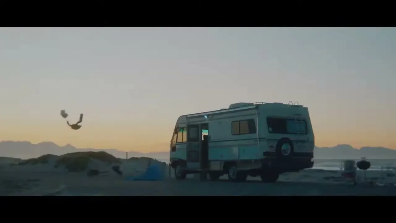A young boy in a blue shirt and shorts jumps off the side of an RV parked on a sandy beach at sunset.