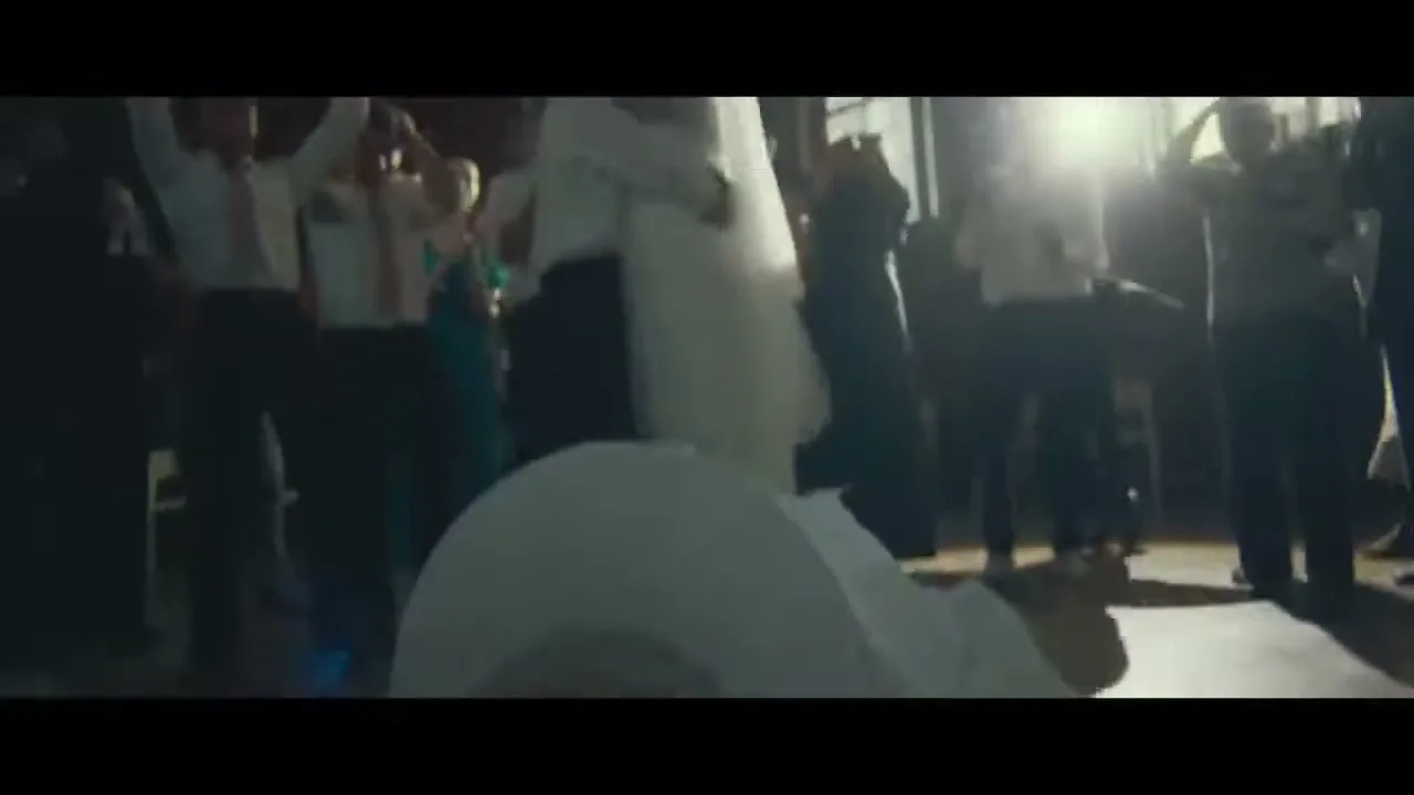 A newlywed couple, the bride in a white dress and the groom in a suit, dance energetically at their wedding reception, surrounded by cheering guests.