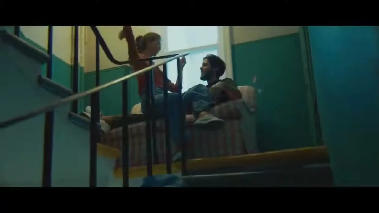 A young couple, a woman in overalls and a man in a t-shirt, sit on a landing in a stairwell, talking and laughing.