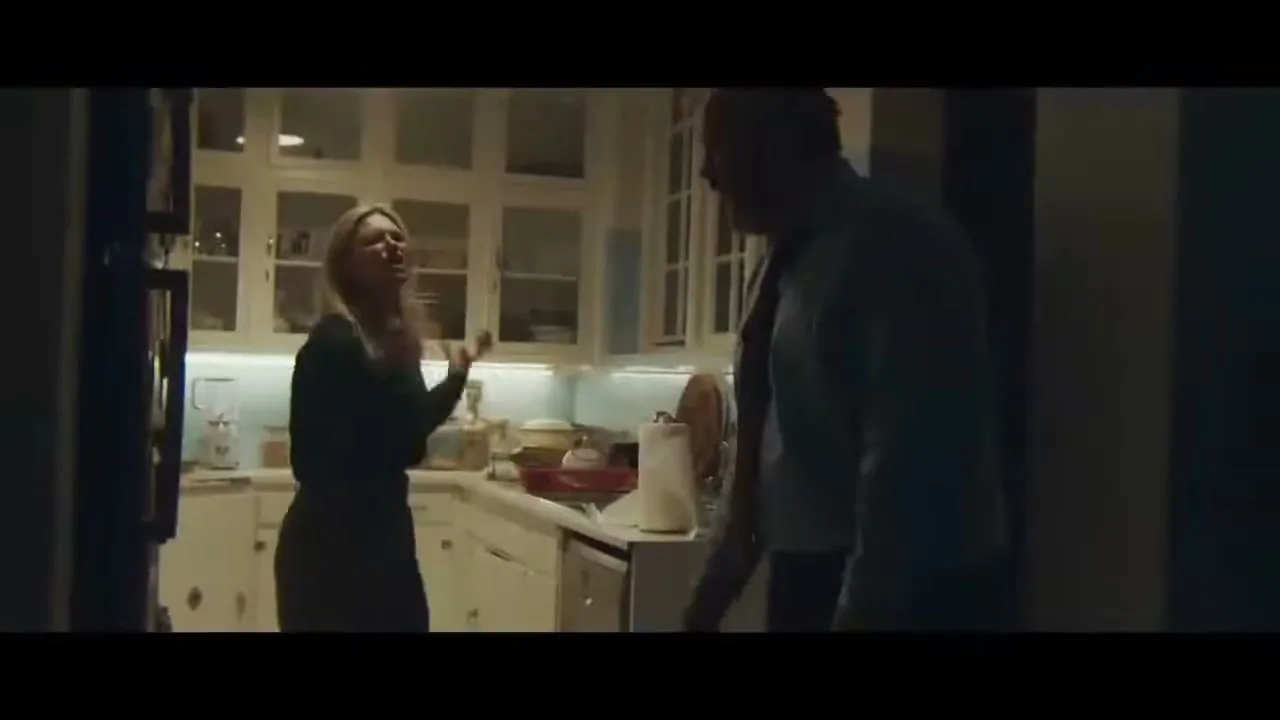 A blonde woman in a dark top and a man in a blue shirt argue in a dimly lit kitchen.