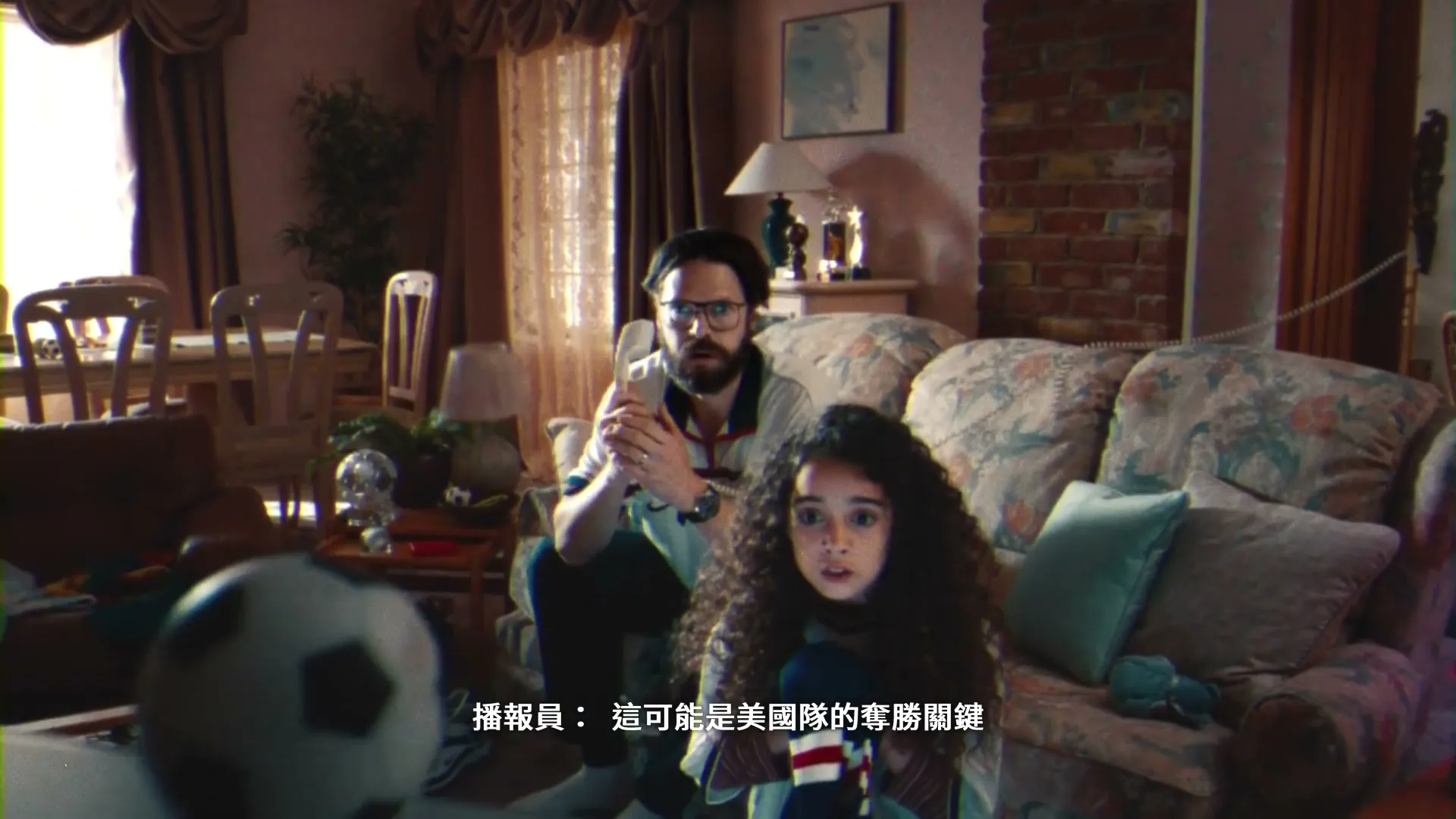 A man with a beard and glasses talks on a landline phone while a young girl with curly hair watches intently, both sitting in a cluttered living room.