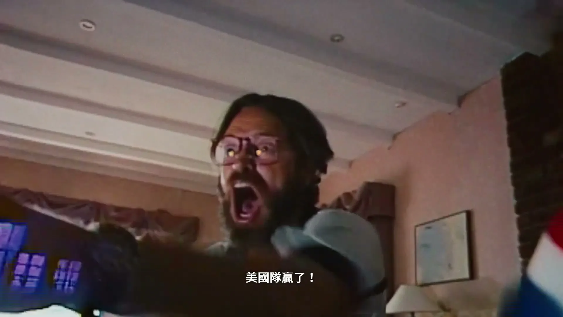 A man with a beard and glasses screams excitedly, wearing a light blue shirt, in a living room with a lamp and a picture frame visible.