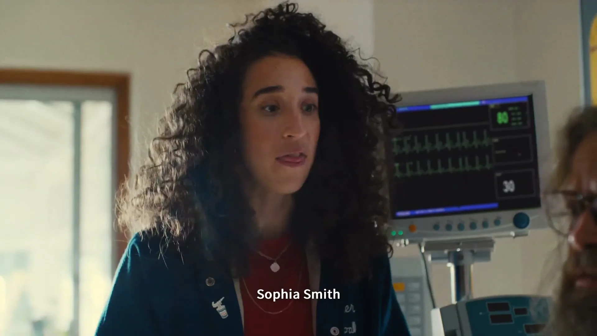 A young woman with curly hair, wearing a red shirt and blue jacket, talks to someone off-screen in a hospital room.
