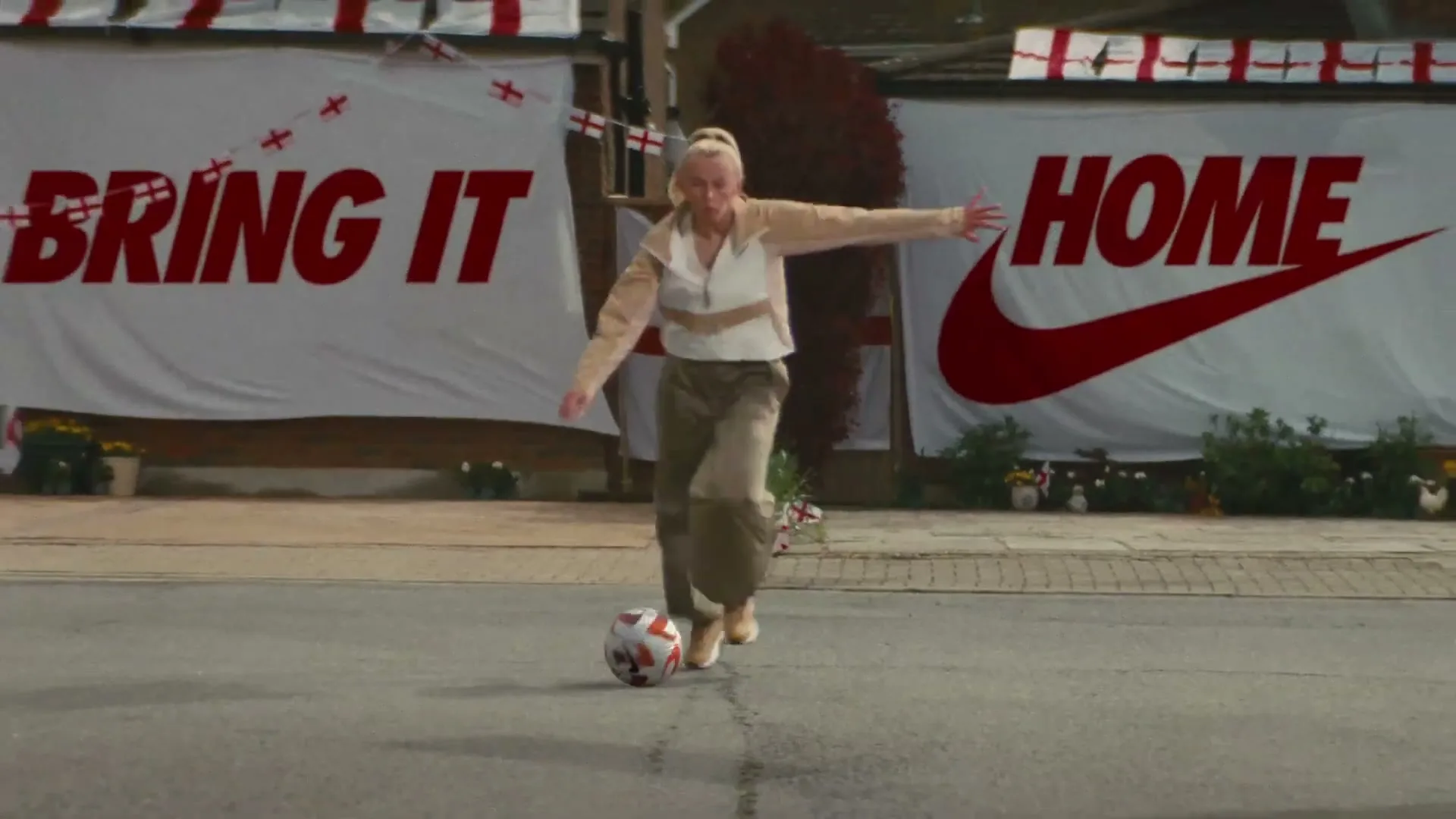A female soccer player in a beige jacket and cargo pants dribbles a soccer ball down a street decorated with England flags and a "Bring It Home" banner.