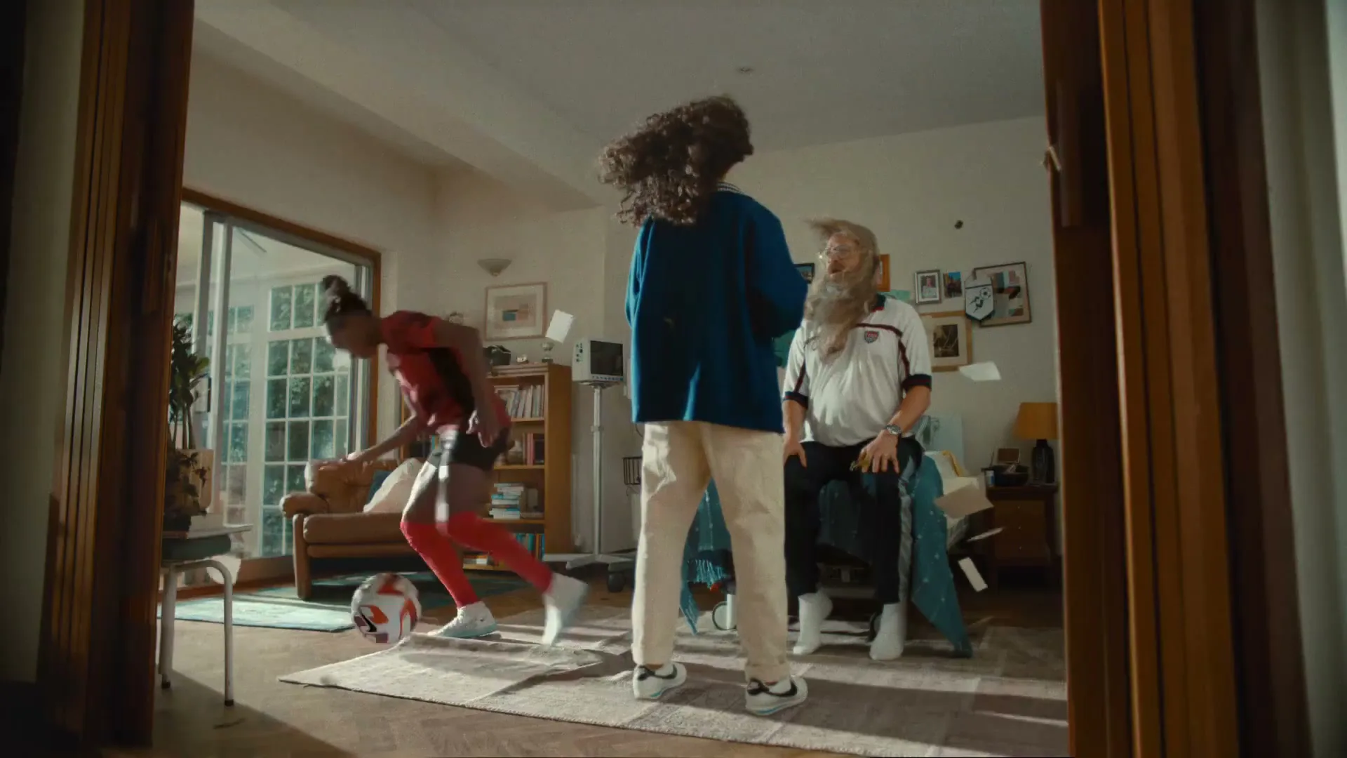 A female soccer player in a red uniform kicks a soccer ball indoors, while two other people watch, one with a surprised expression.