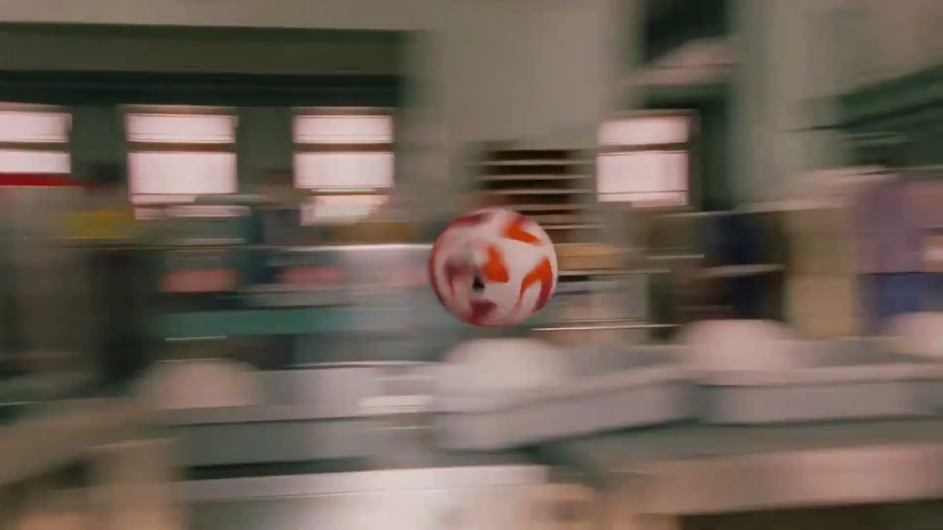 A female soccer player in a blue uniform dribbles a soccer ball through a busy mailroom, surrounded by boxes and mail carts.