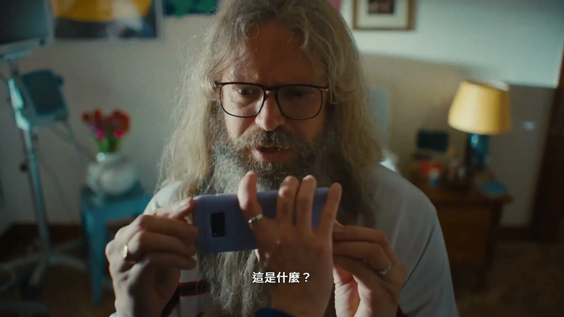 A man with long hair and a beard holds a phone horizontally, looking at it with a surprised expression in a cluttered room.