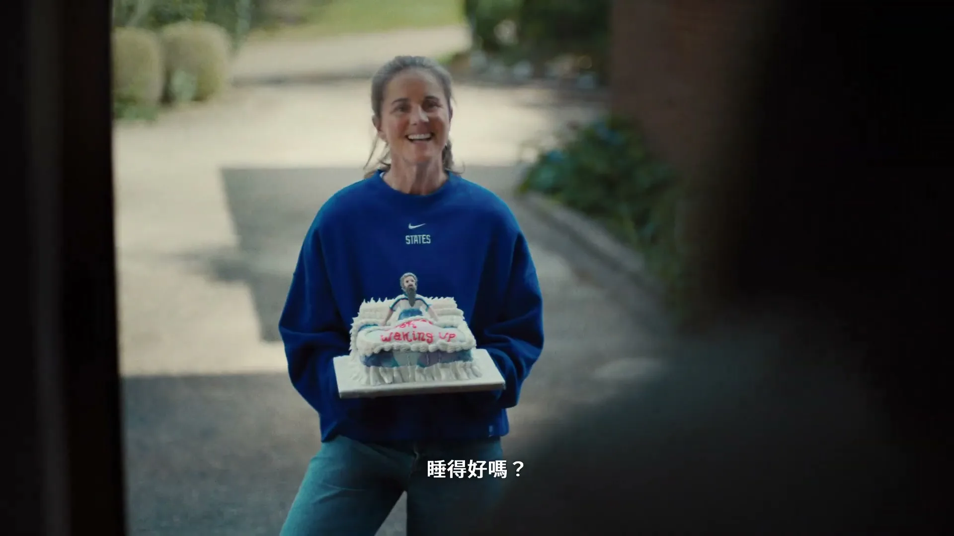 A smiling woman in a blue sweatshirt holds a custom cake with a sleeping man on it, standing outside a house.